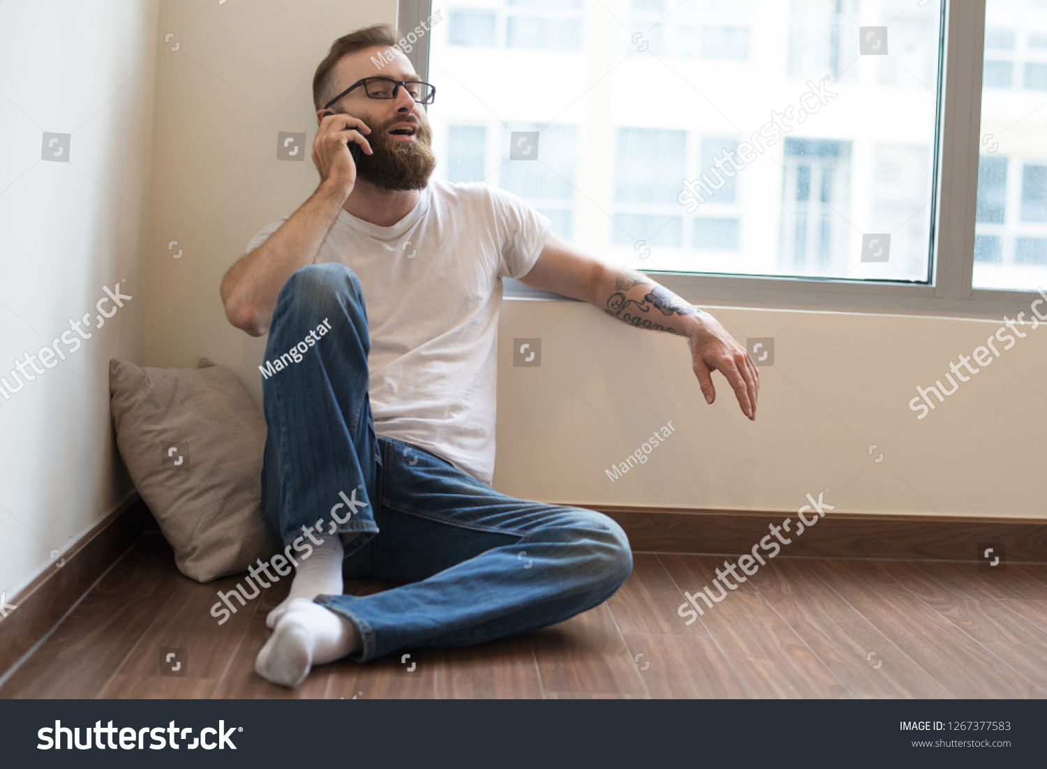 Confident handsome young man in glasses talking on phone. Content bearded guy sitting on floor and expressing his opinion while communicating on phone. Conversation concept #1267377583