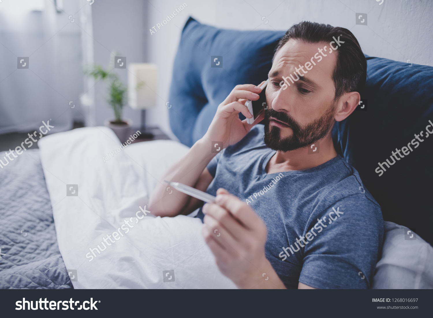 portrait of sick man holding thermometer and calling a doctor while sitting on the bed #1268016697