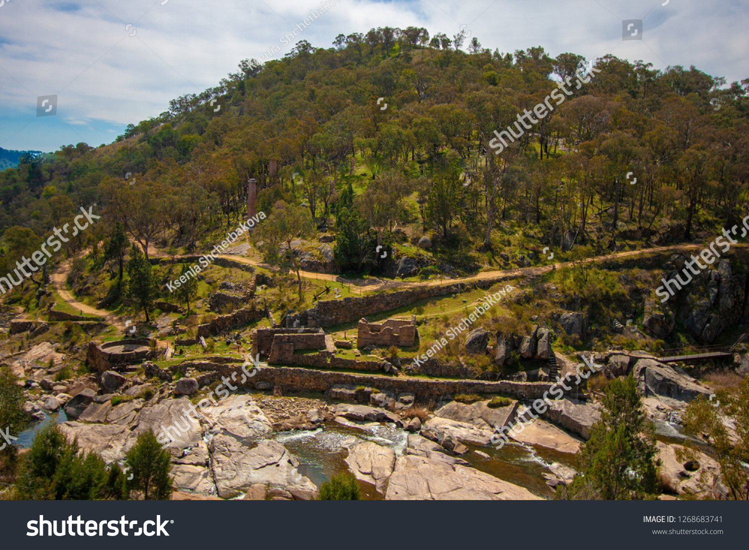 Narrow small mountain river at the foot of a round green hill. At the foot of the hill are the ruins of a gold factory. Australia. #1268683741
