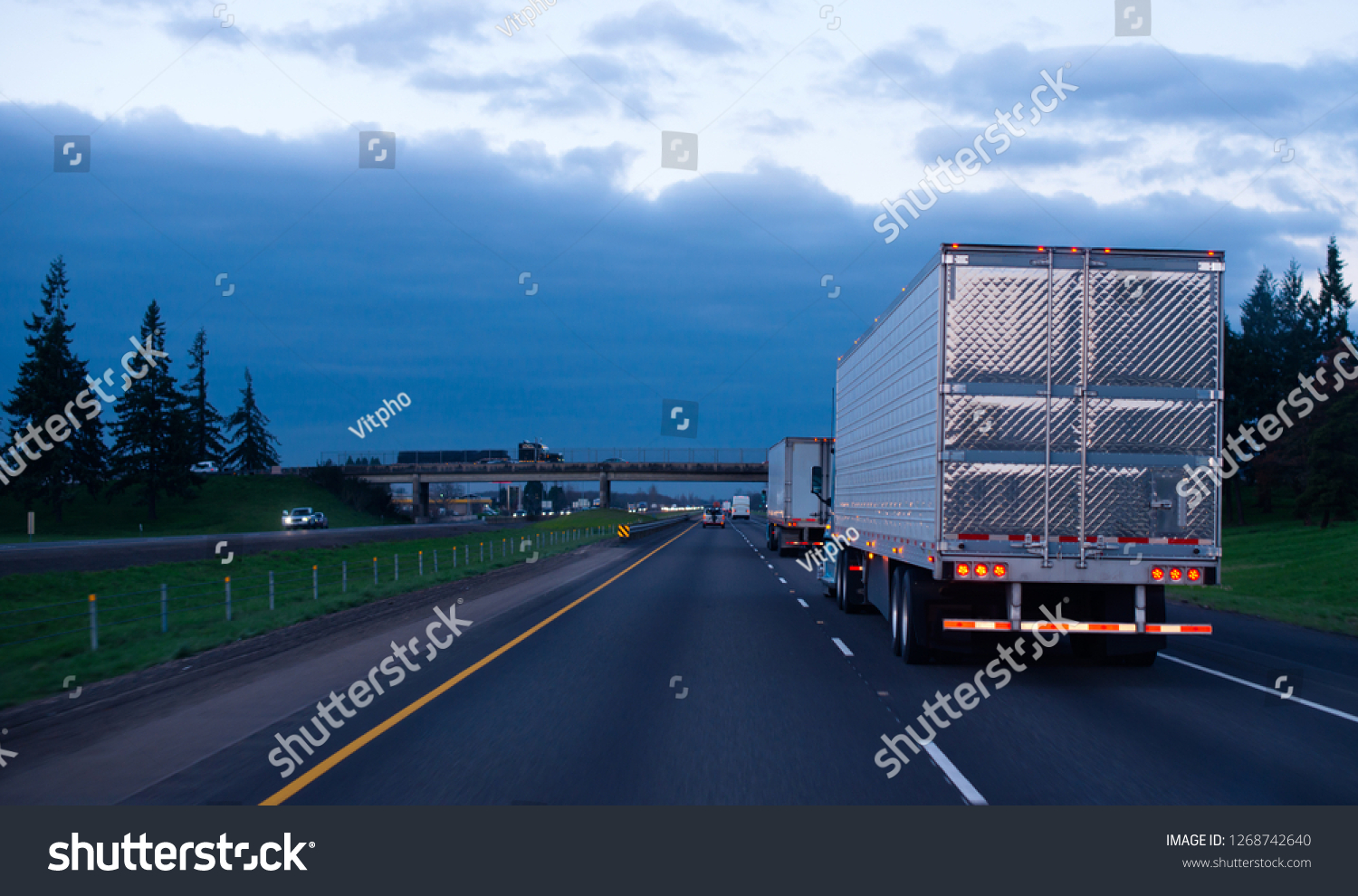 The convoy of semi trucks with reefer trailers on flat like an arrow evening road with lights on and reflection of light on a shiny trailer stainless steel doors. Trucks driving on a divided highway #1268742640