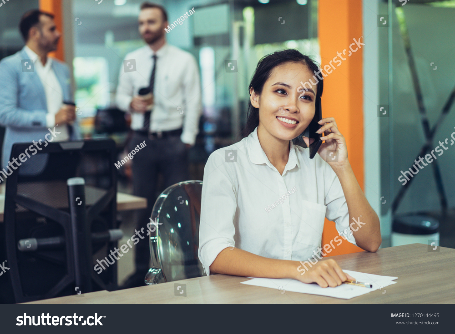 Smiling young Asian business woman talking on phone and working at desk in office. Two coworkers relaxing and standing in background. Management and communication concept. #1270144495