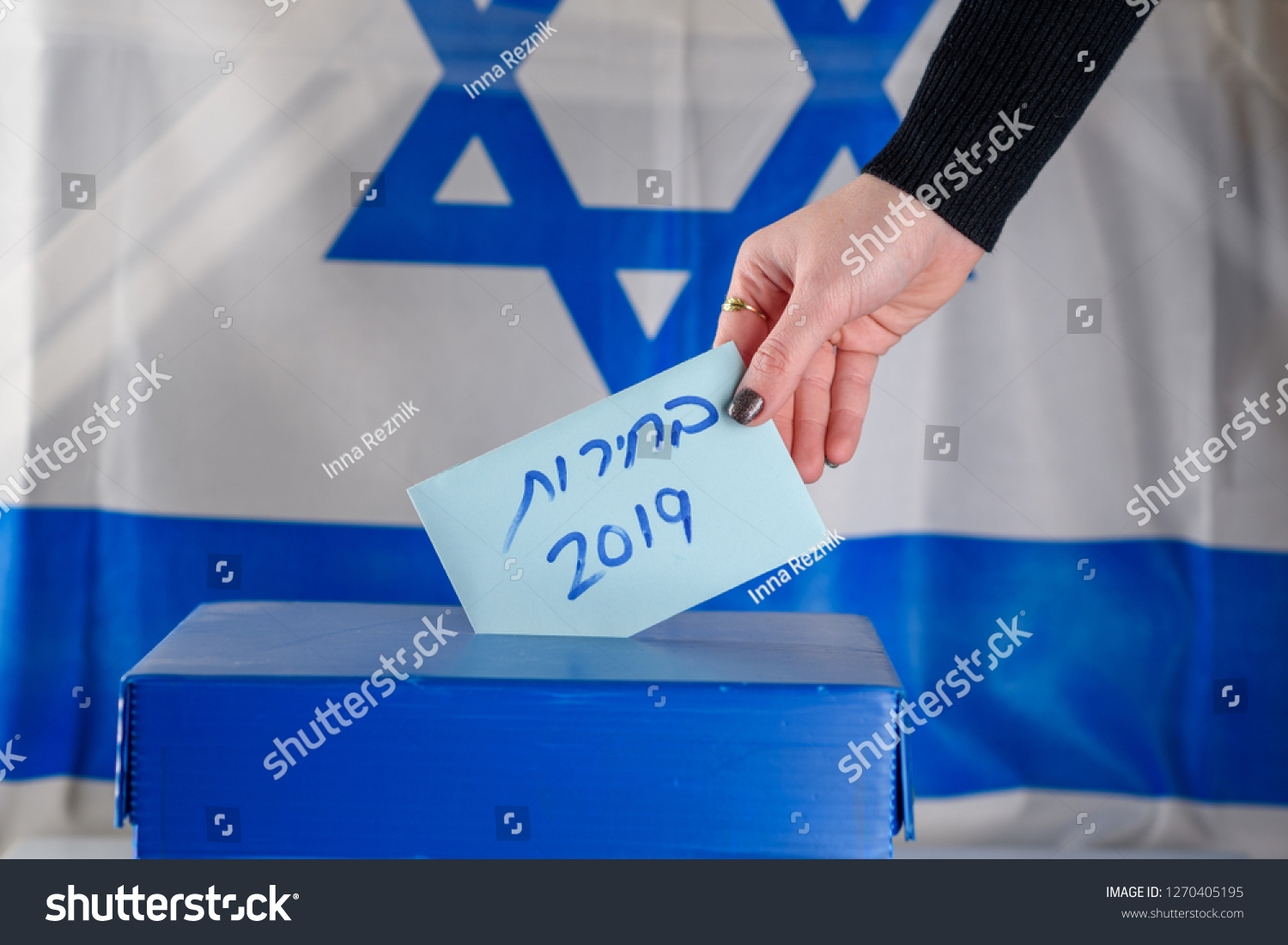 Election in Israel - voting at the ballot box. The hand of woman putting her vote in the ballot box. Israeli Flag on background. Hebrew text Elections 2019. #1270405195
