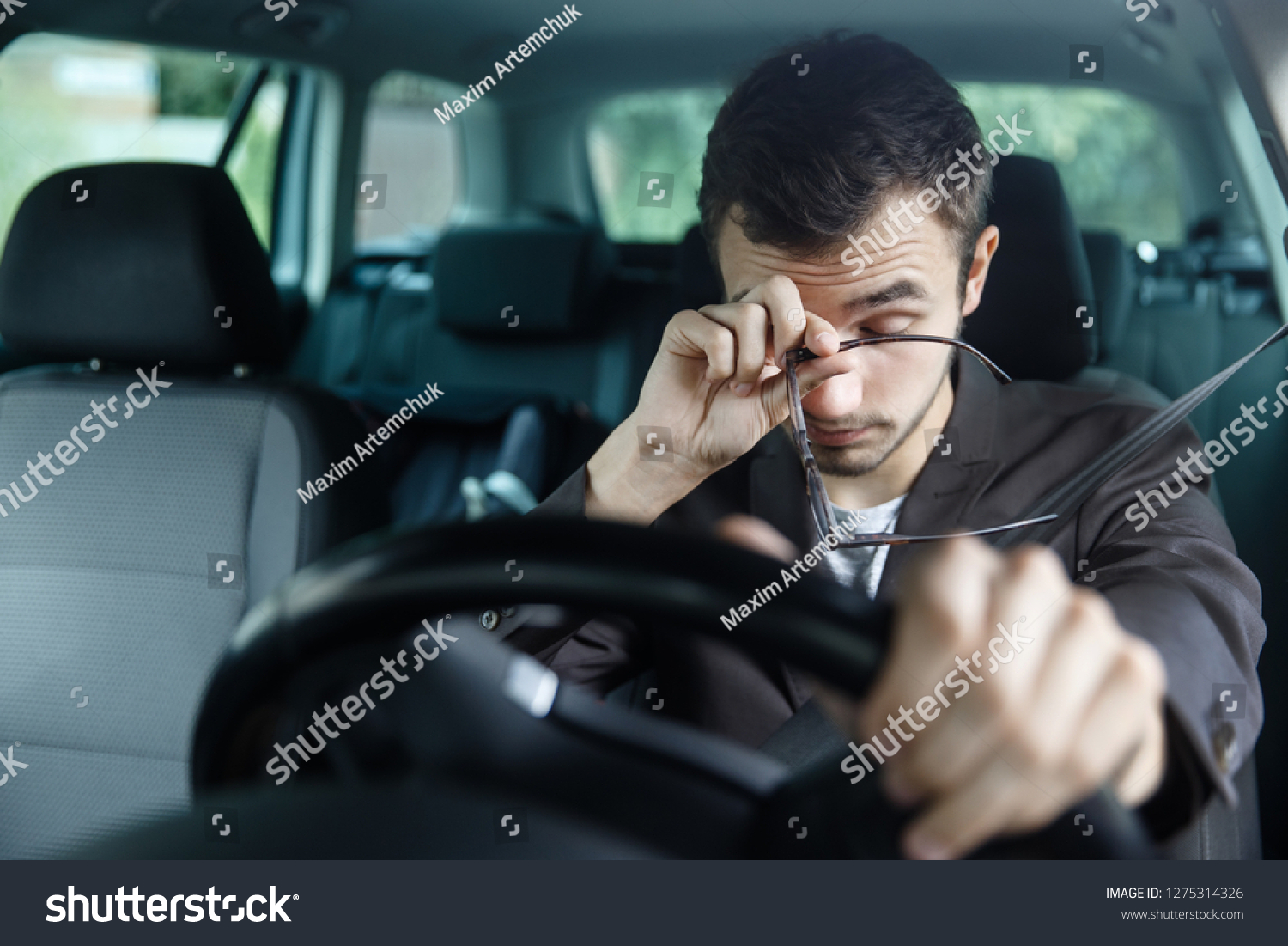 Sleepy young man rubs his eyes with his right hand. His left hand is on the steering wheel. He is sitting at his car. Road safety concept. #1275314326