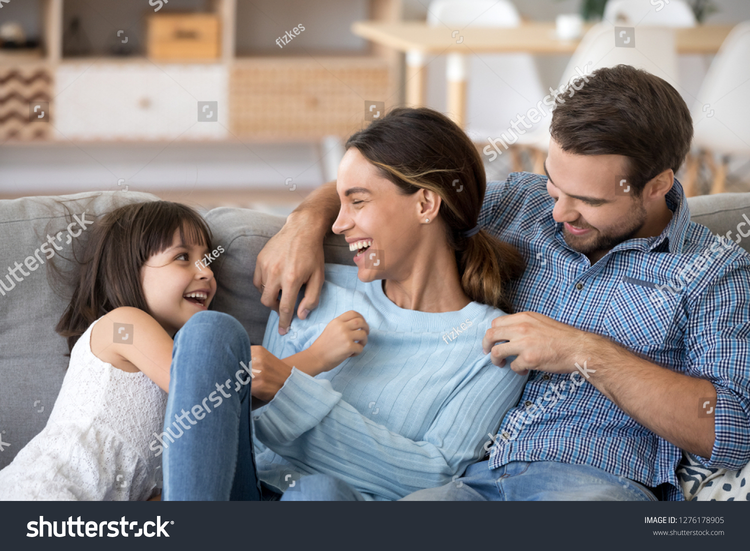 Cute kid daughter and dad tickling mom having fun good time playing together at home, happy parents and little child girl enjoying funny activity and communication, family laughing relaxing on sofa #1276178905