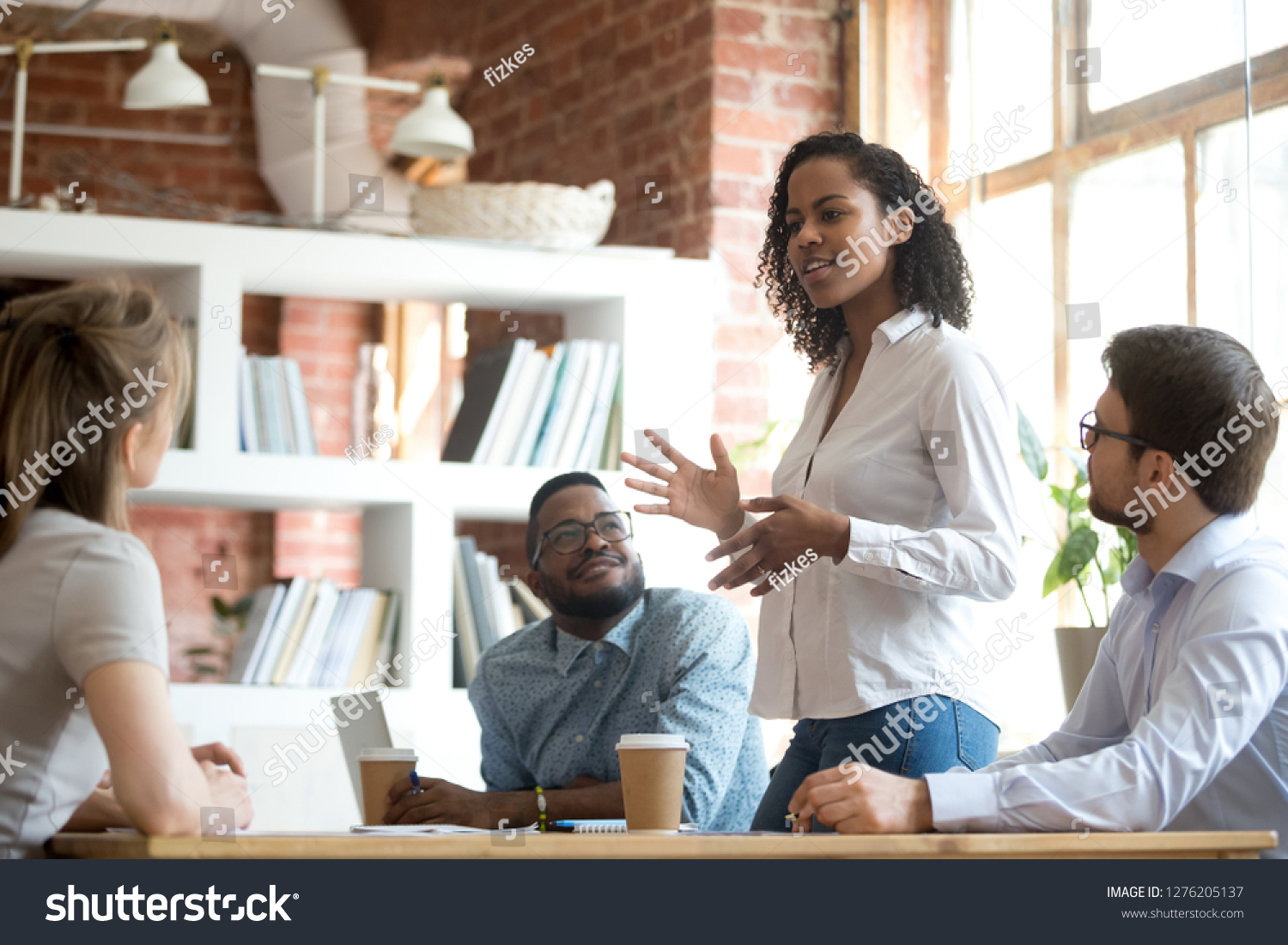 Ambitious smart african black female employee speaking at diverse meeting share creative idea opinion at group briefing while jealous envious skeptical male coworkers looking listening to colleague #1276205137