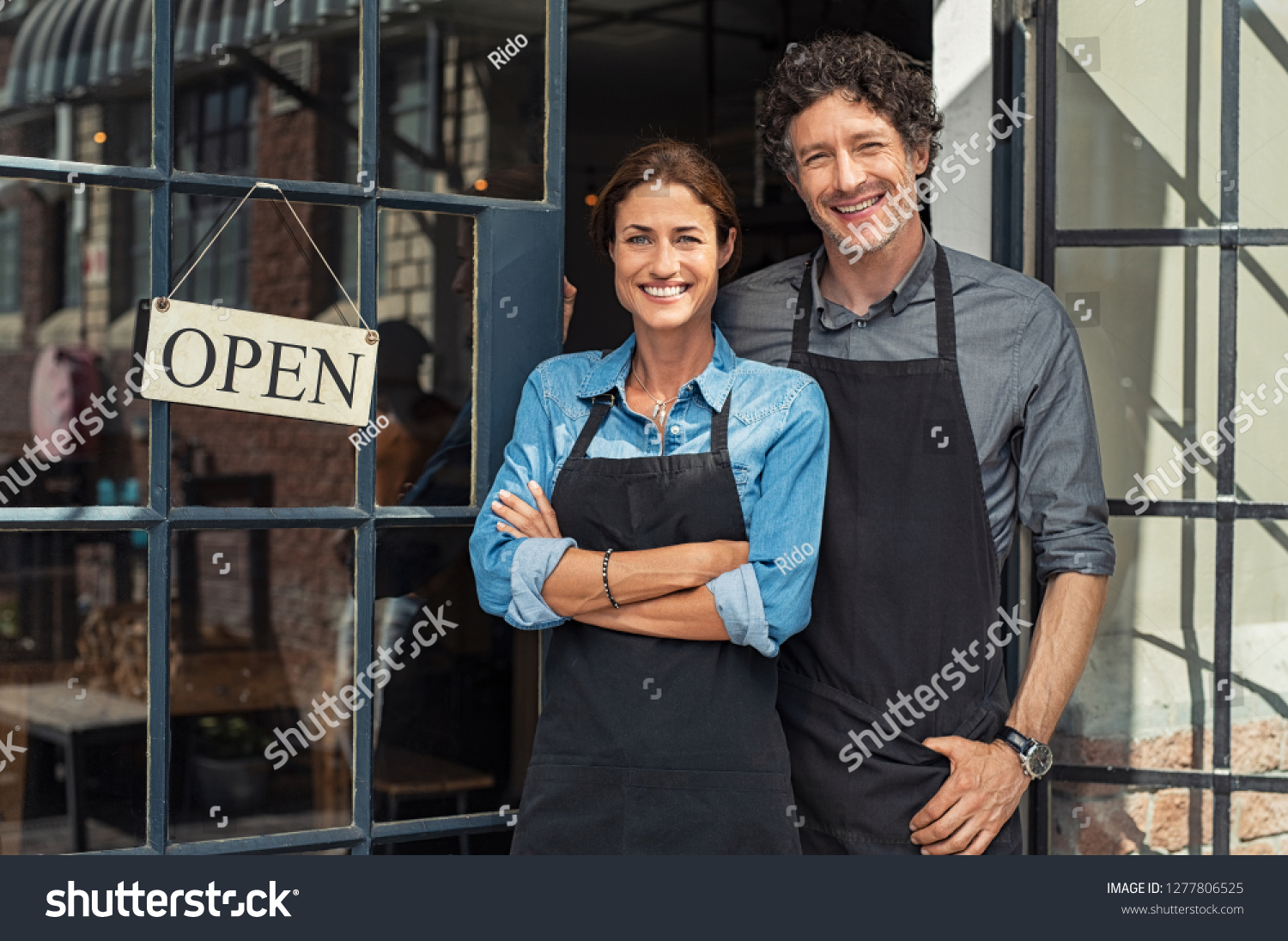 Two cheerful small business owners smiling and looking at camera while standing at entrance door. Happy mature man and mid woman at entrance of newly opened restaurant with open sign board. #1277806525