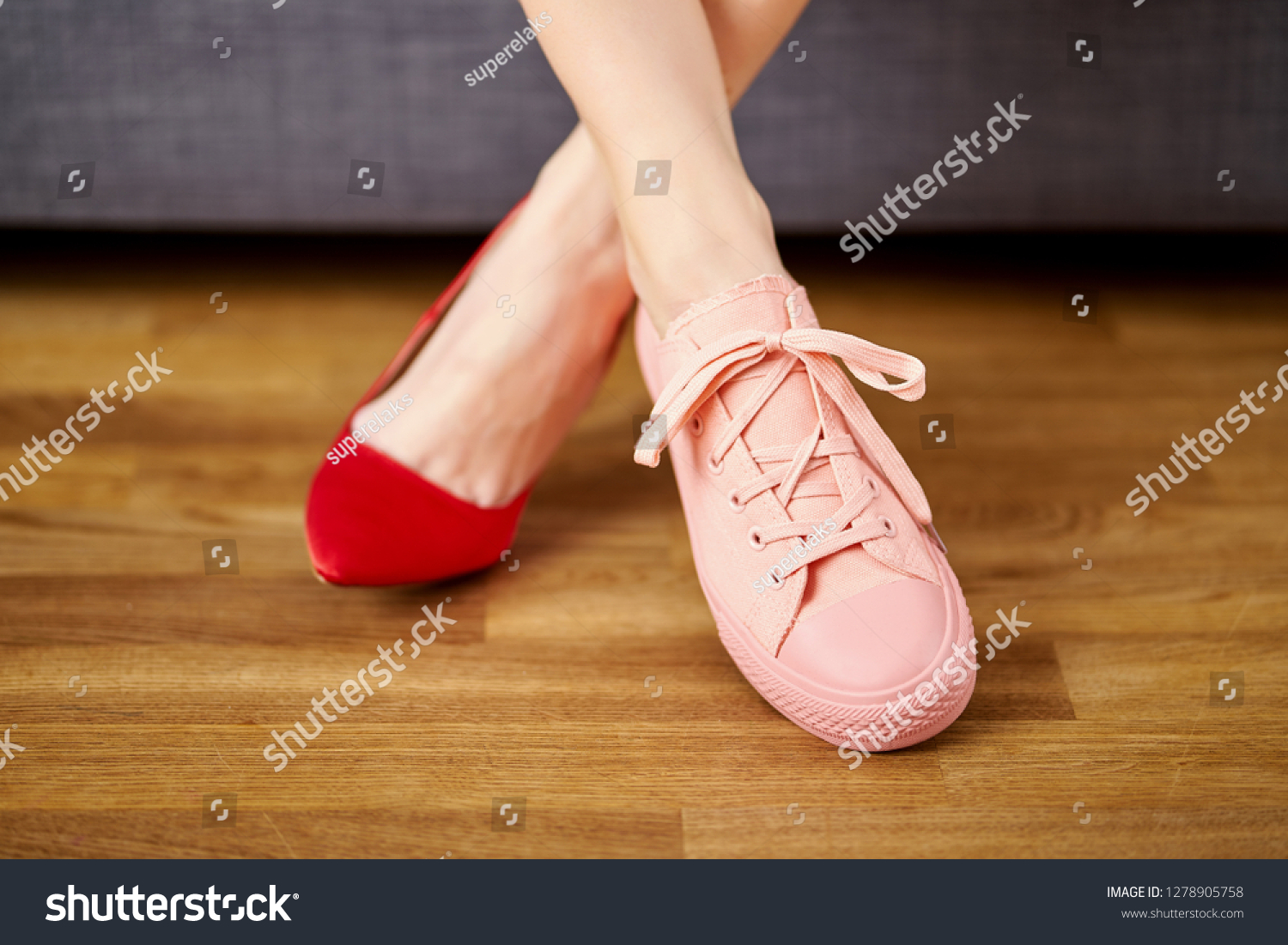 Slim woman's legs in two different shoes with red high heels and coral sneakers on gray couch background. #1278905758