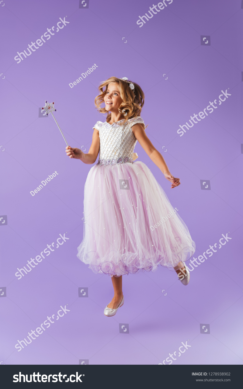 Full length portrait of a smiling pretty girl dressed in a princess dress isolated over violet background, holding magic wand, jumping #1278938902