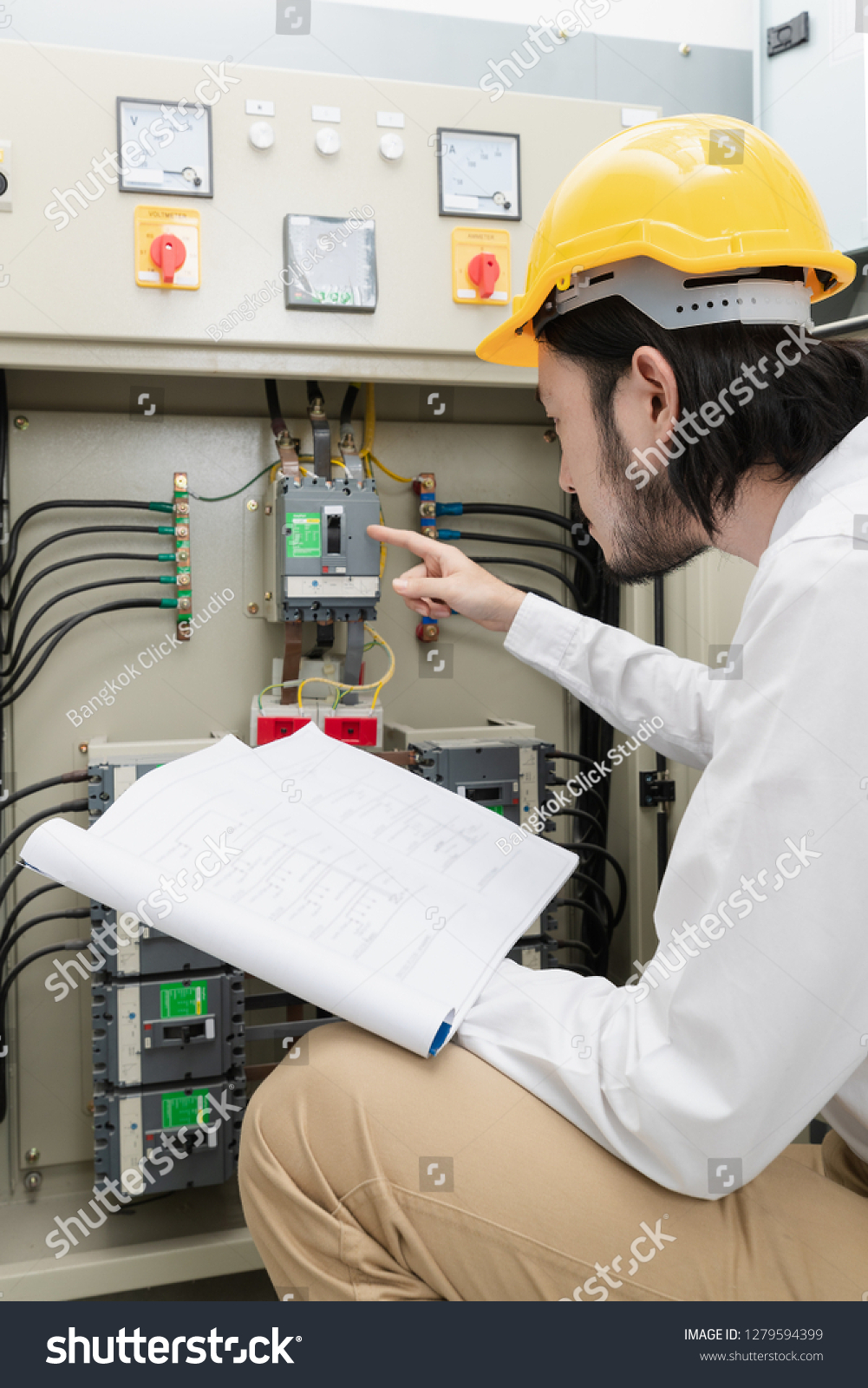 Close up of young Asian electric engineer in beige pant and white shirt, wear yellow helmet, squat near control panel, working instruction in hand, follow up his routine daily checking #1279594399