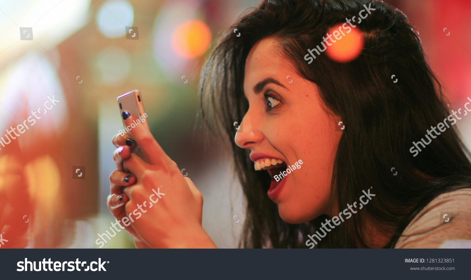 Candid surprise reaction of girl in front of cellphone reading text message smiling and laughing at night at coffee shop #1281323851