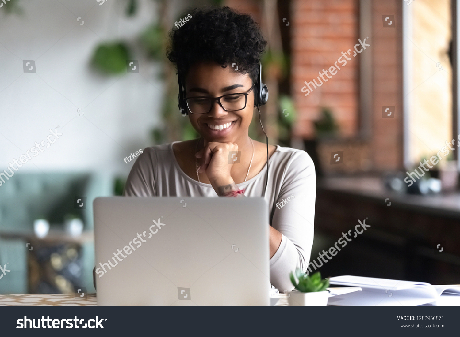 Black woman smart student girl sitting at table in university cafe alone wearing glasses looking at computer screen using headphones listening online lecture improve language skills having good mood #1282956871