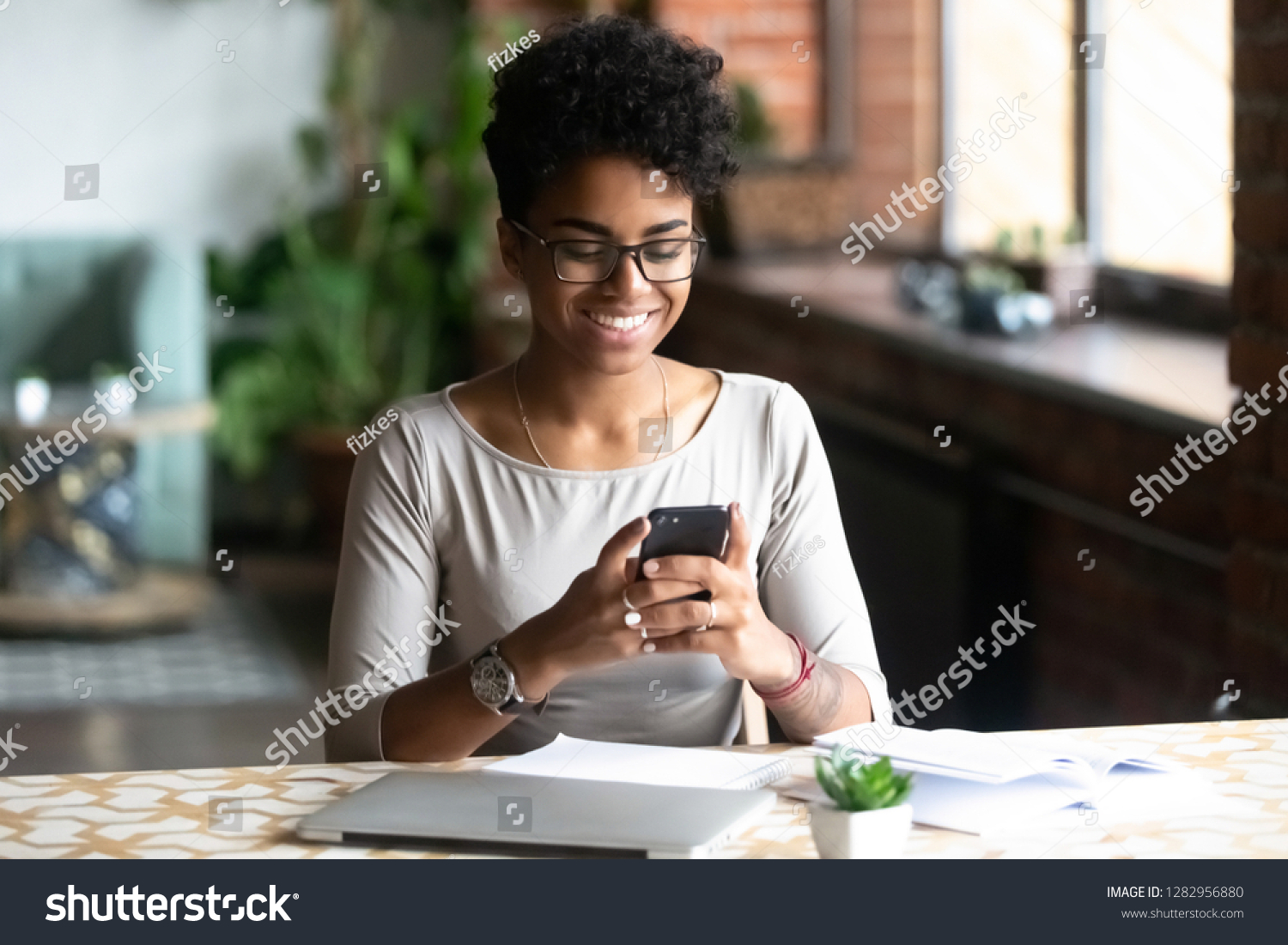 Cheerful african student black woman sitting at table studying using laptop reading a book, take a break holding mobile phone surfing internet received message from friend chatting about weekend plans #1282956880