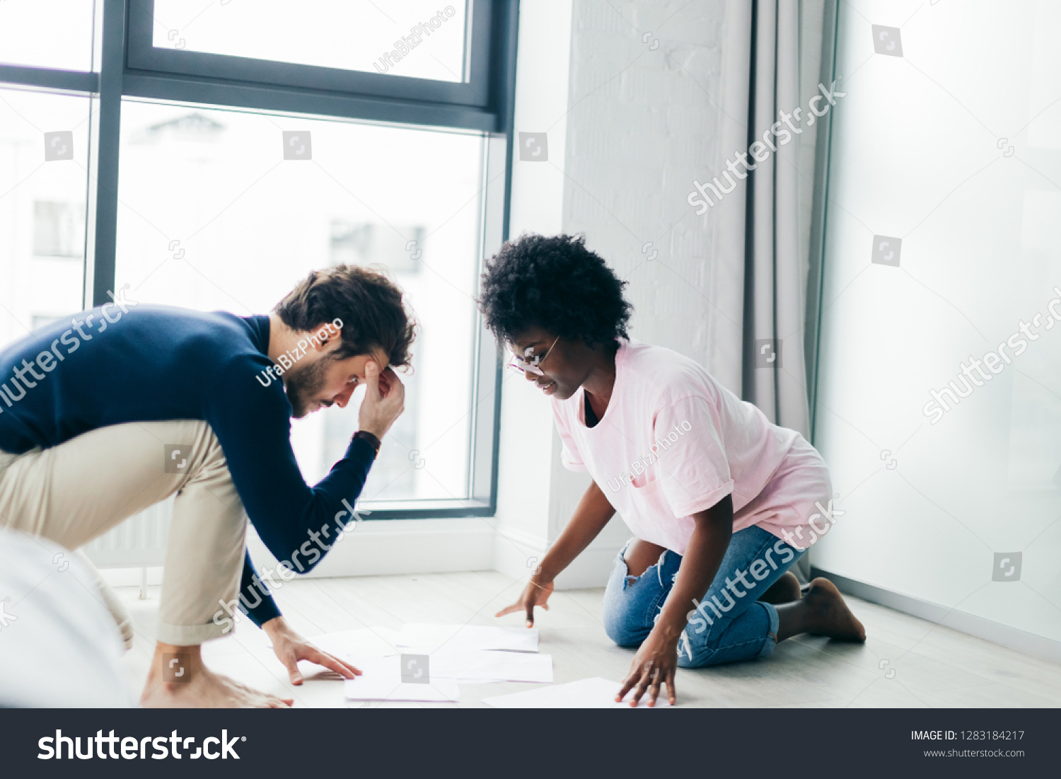 Focused multicultural small business partners develop future project, kneeling on floor in living room, looking at papers, thinking over how to make investments. #1283184217