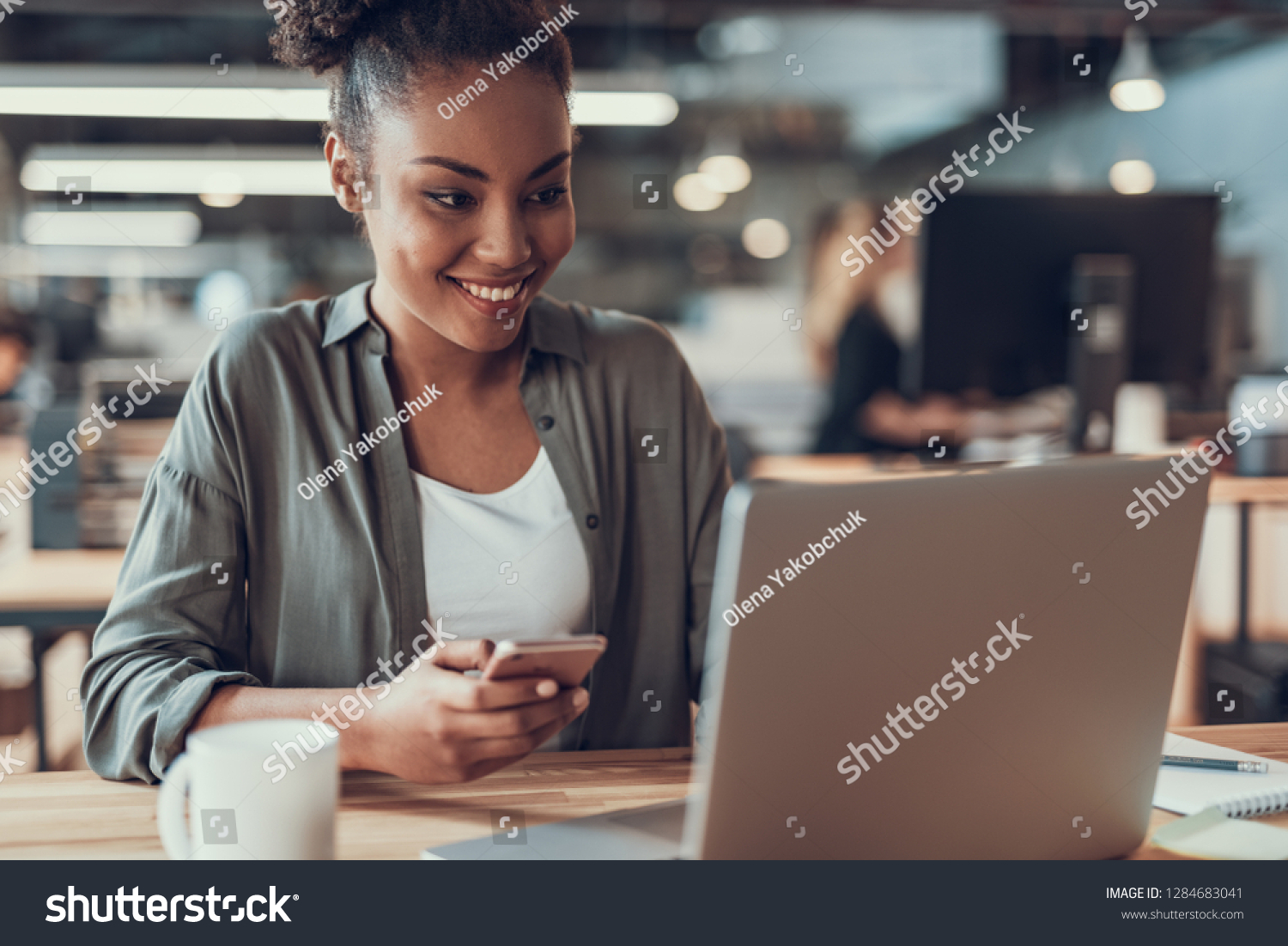 Portrait of charming young lady holding smartphone while looking at notebook display. She is sitting at wooden table and smiling #1284683041