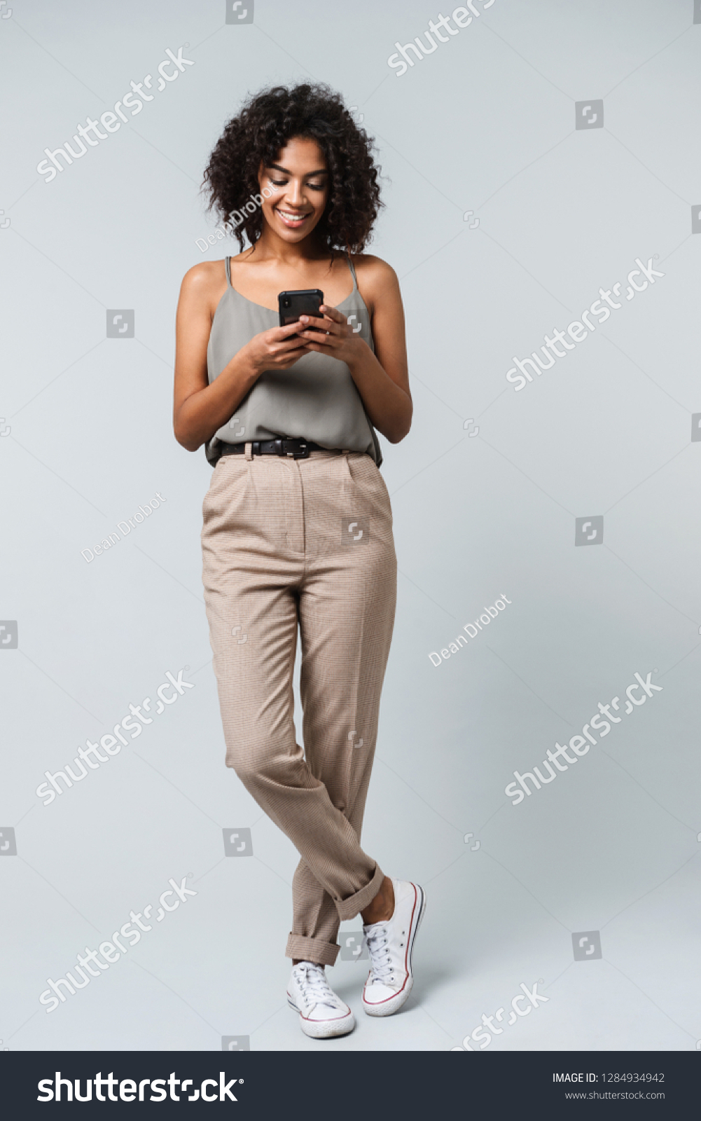 Full length of a happy young african woman casually dressed standing isolated over gray background, holding mobile phone #1284934942