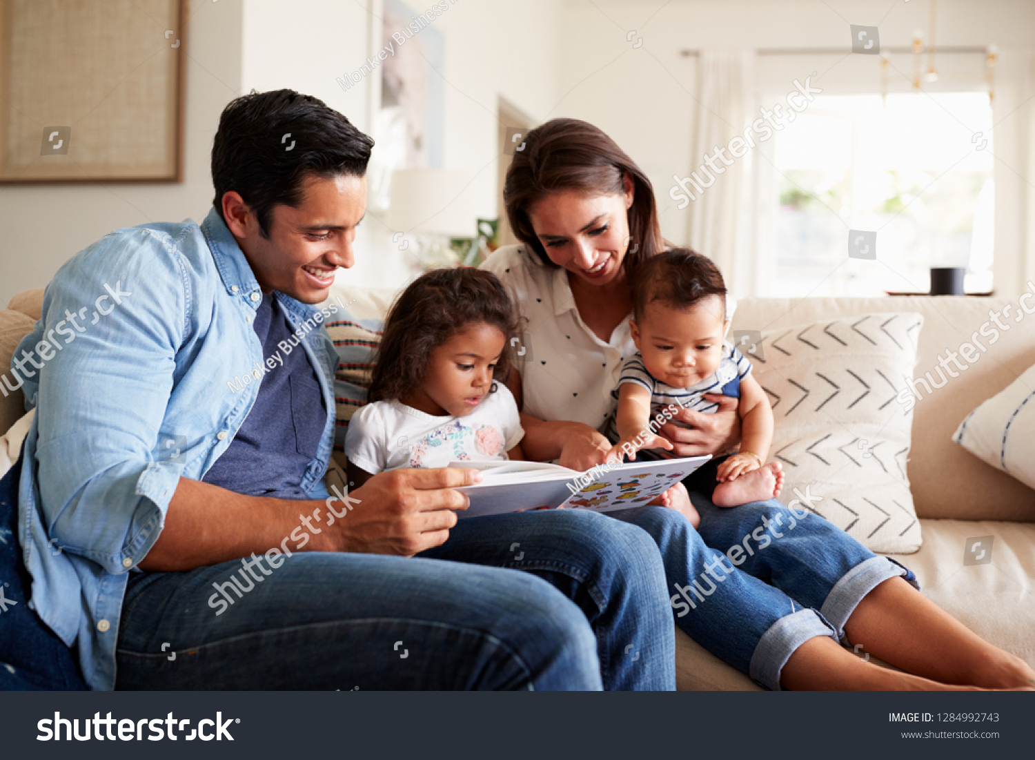 Young Hispanic family of four sitting on the sofa reading a book together in their living room #1284992743