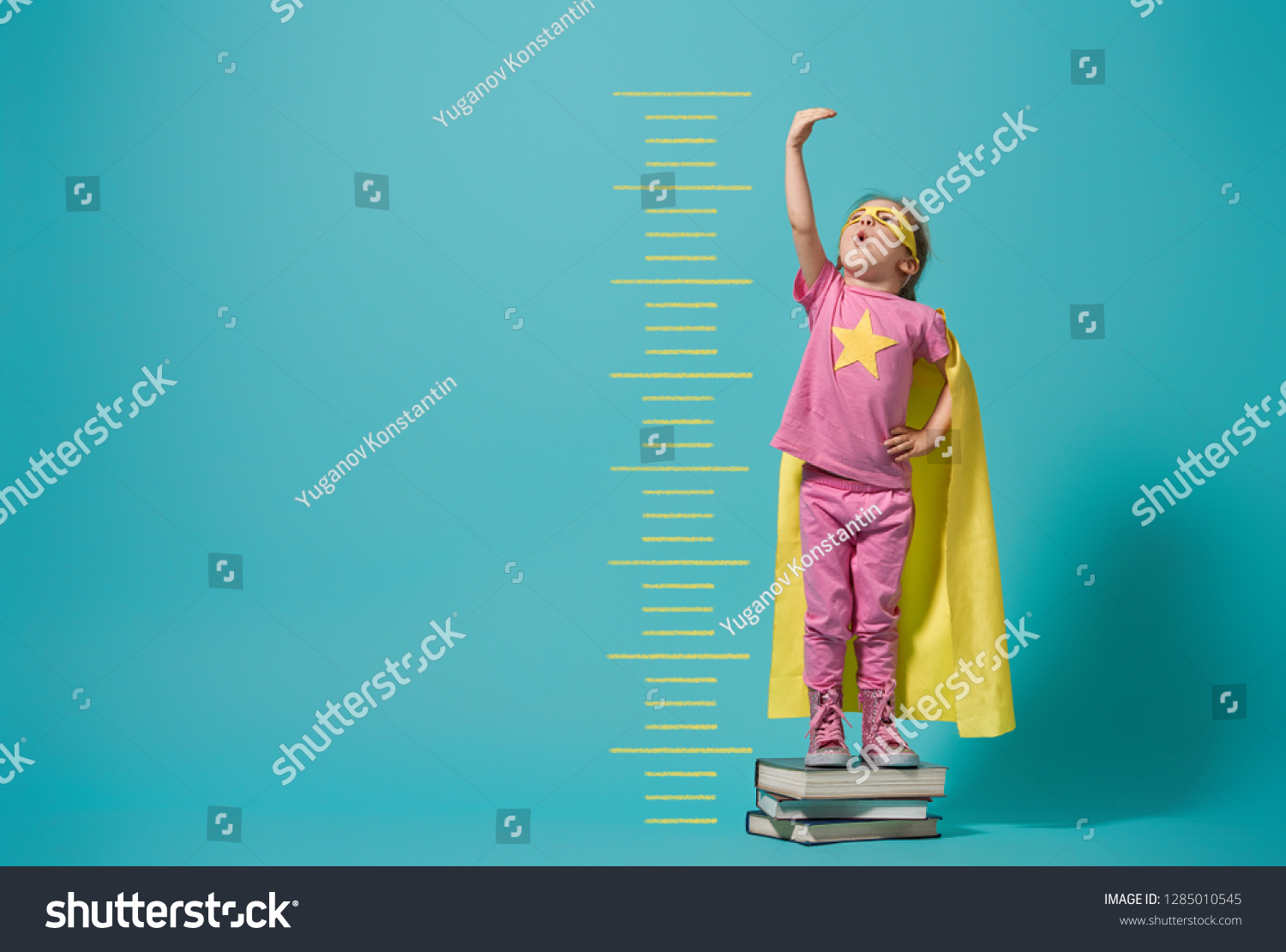 Little child playing superhero. Kid measures the growth on the background of bright blue wall. Girl power concept. Yellow, pink and  turquoise colors. #1285010545