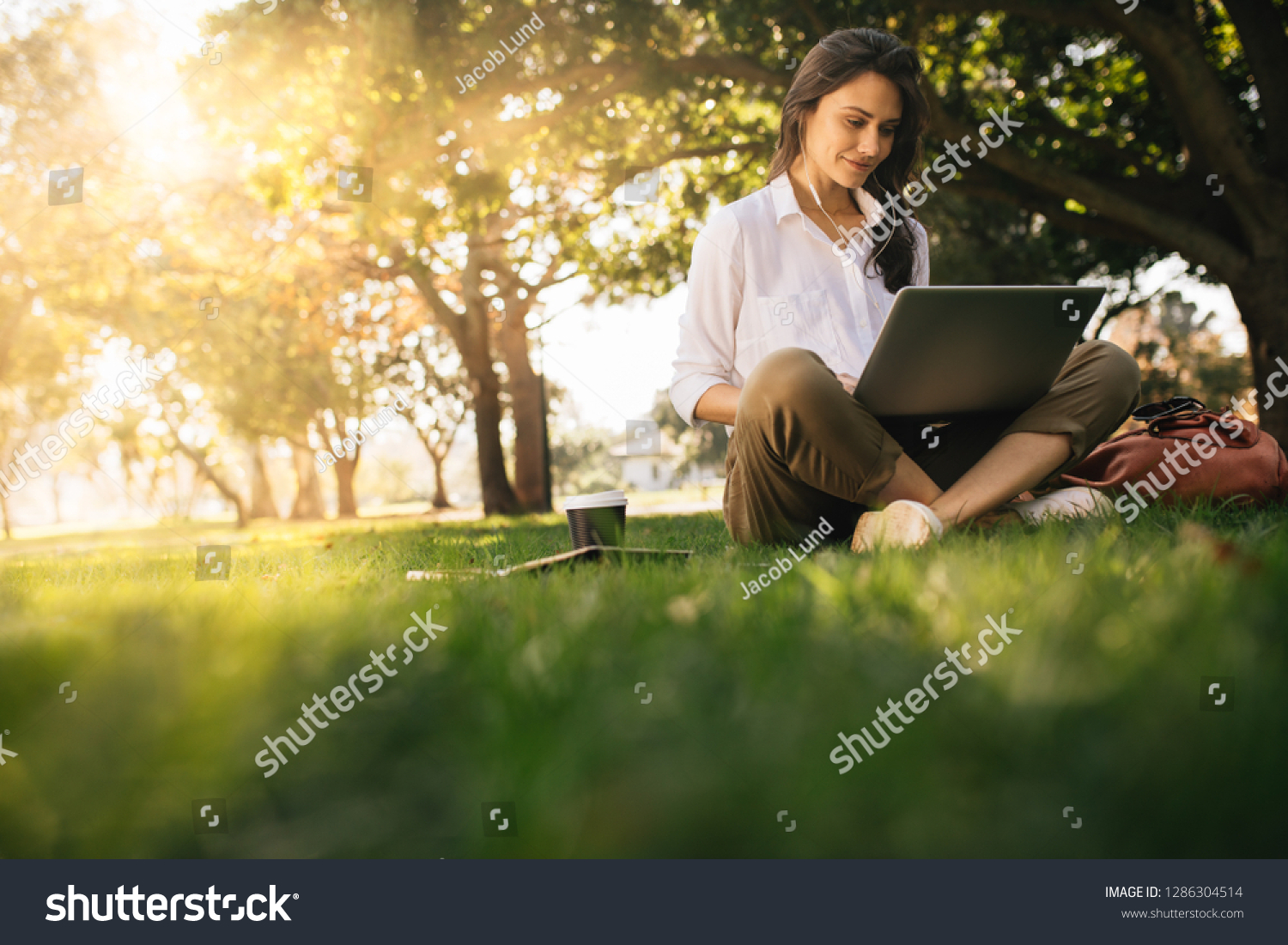 Woman sitting on grass at park working on laptop. Female wearing earphones using laptop while sitting under a tree at park with bright sunlight from behind. #1286304514