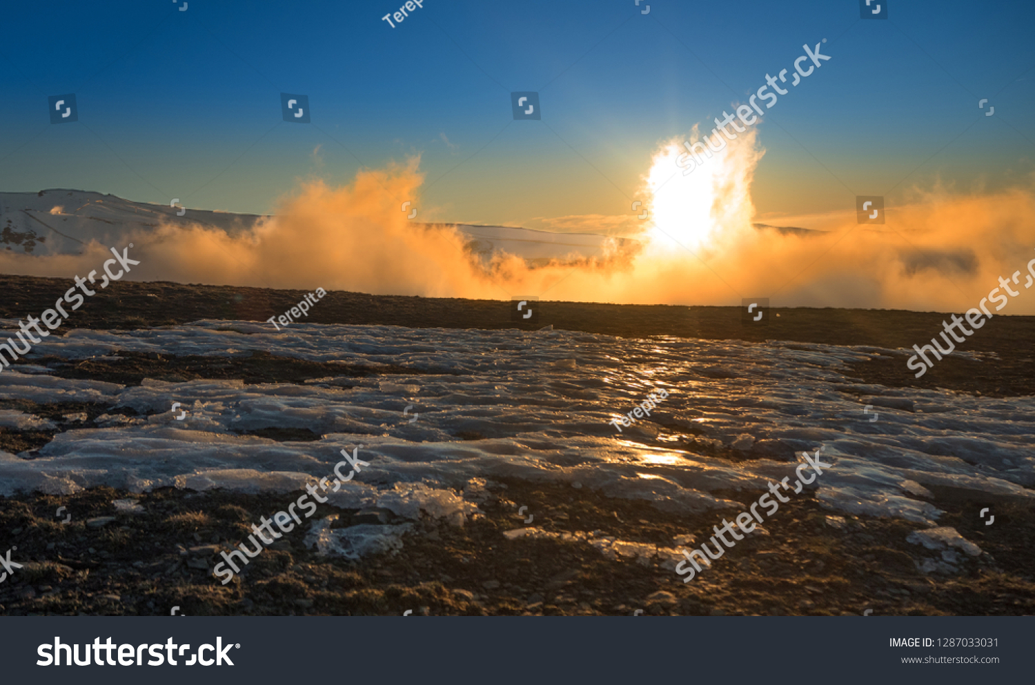Close-up of brown land soil with snow and ice followed by a thick white mist that  traverses the orange light of the sun that is behind a snowy white mountain under the dark blue sky #1287033031