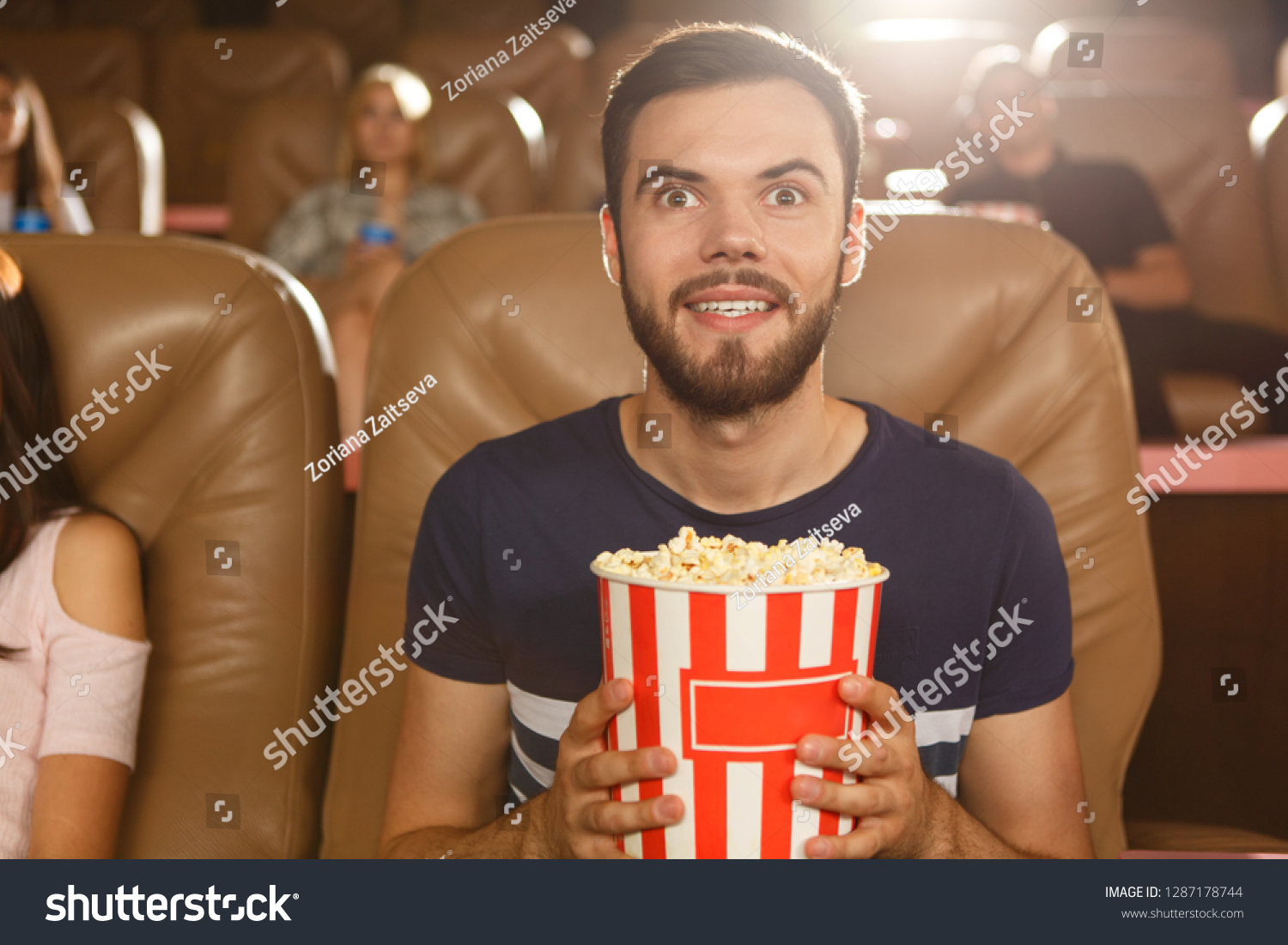 Excited male viewer keeping popcorn in hands and watching interesting film in cinema. Young bearded man wearing shirt expecting final of movie. Concept of entertainment and leisure. #1287178744