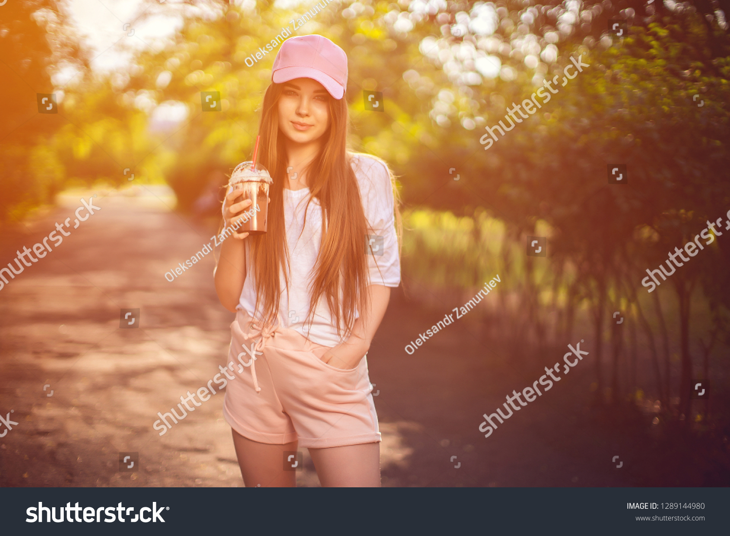 Pretty confident brunette in cap and summer outfit holding chocolate drink in cup looking at camera with smile in nature #1289144980