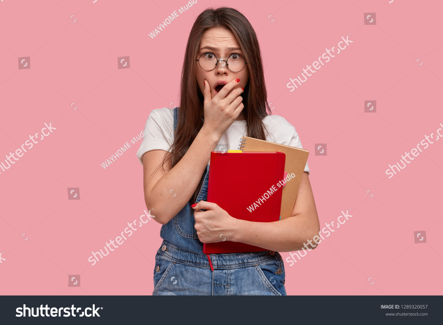 Emotive freckled woman covers mouth, afraids of scarying scene, carries notepad, wears casual white t shirt and overalls, poses against pink background. People, studying and emotions concept #1289320057