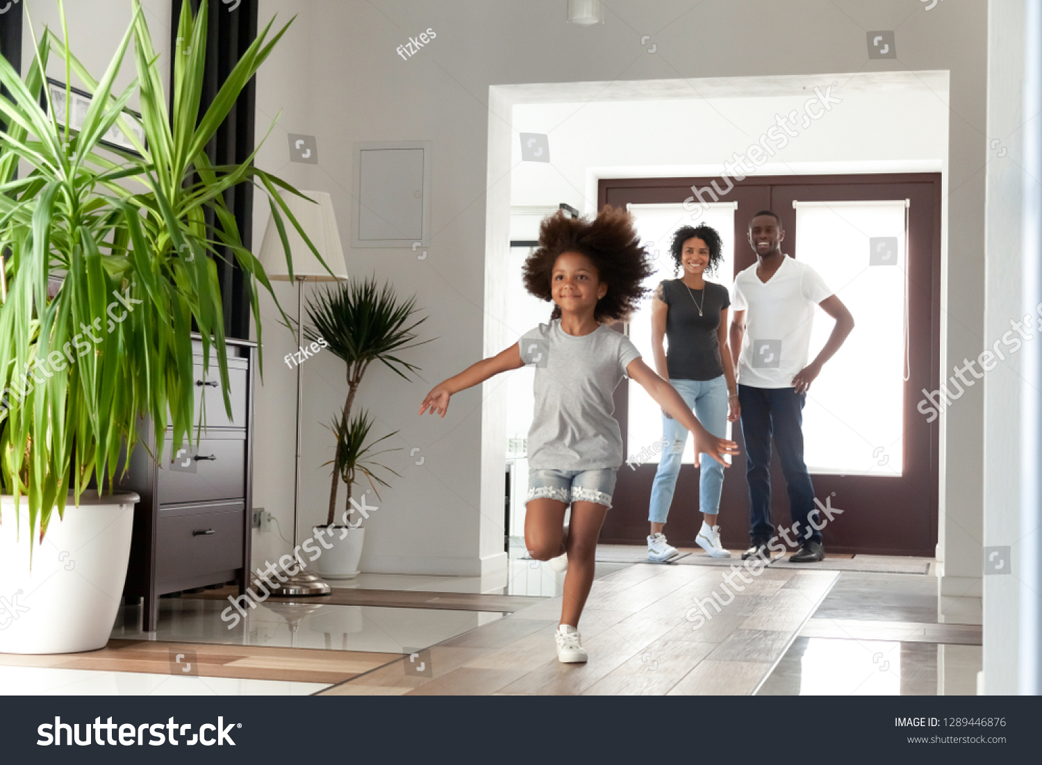 Excited little funny african girl running exploring big modern house moving in, happy black parents and kid daughter coming into new home, cute mixed race child having fun in hallway, family mortgage #1289446876