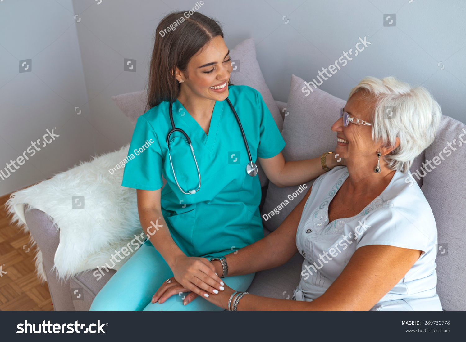 Young caring lovely caregiver and happy ward. Image of caregiver and senior resting in the living room. Smiling caregiver taking care of a happy elderly woman #1289730778