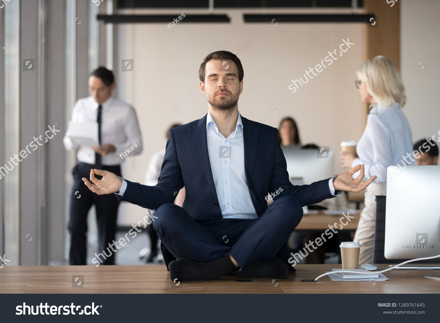 Mindful calm businessman in suit meditating at office sitting in lotus position on work desk, successful ceo executive doing yoga exercise at workplace, peace of mind, no stress free relief concept #1289761645