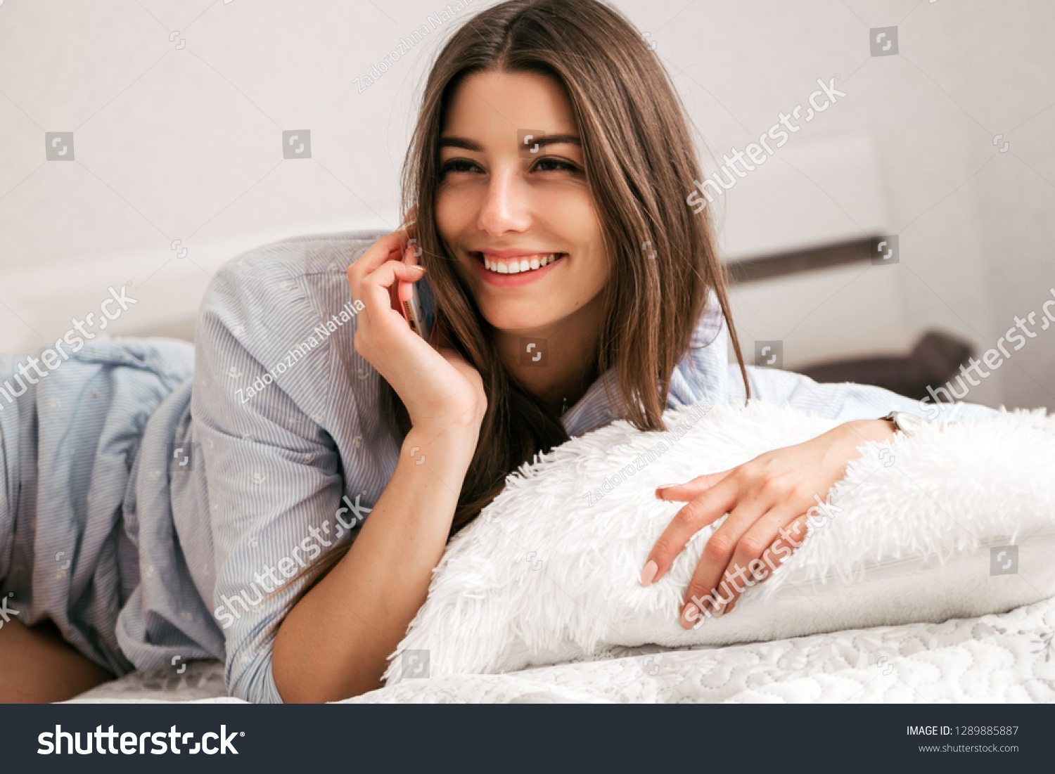 Young smiling woman lying in white bed and using a phone in her bedroom. Happy morning. #1289885887