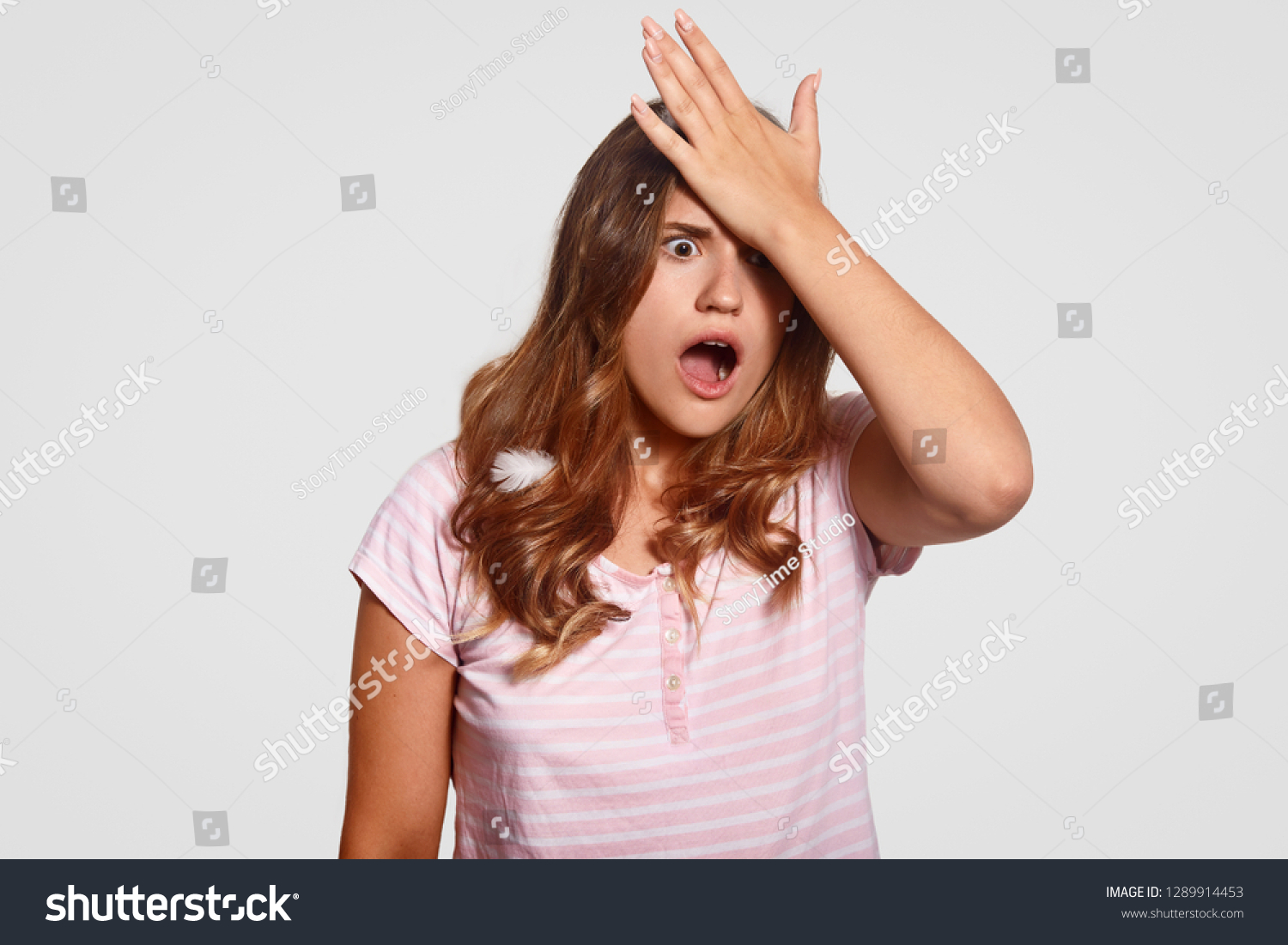 Frustrated shocked woman keeps palm on forehead, forgets about work, wakes up late, dressed in pyjamas, stands against white background, has surprised expression, feathers on hair. Omg concept #1289914453