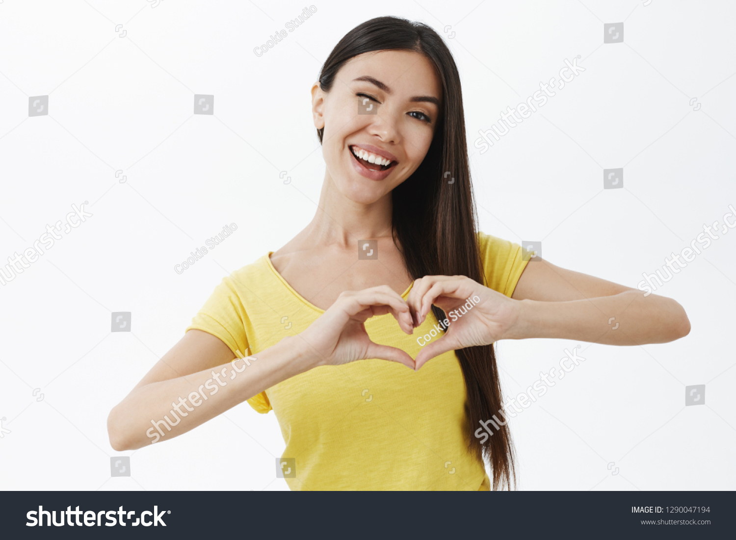 Waist-up shot of cute caring and happy girlfriend with lond dark hair winking joyfully smiling showing heart gesture over chest expressing love and affection being tender and friendly over grey wall #1290047194