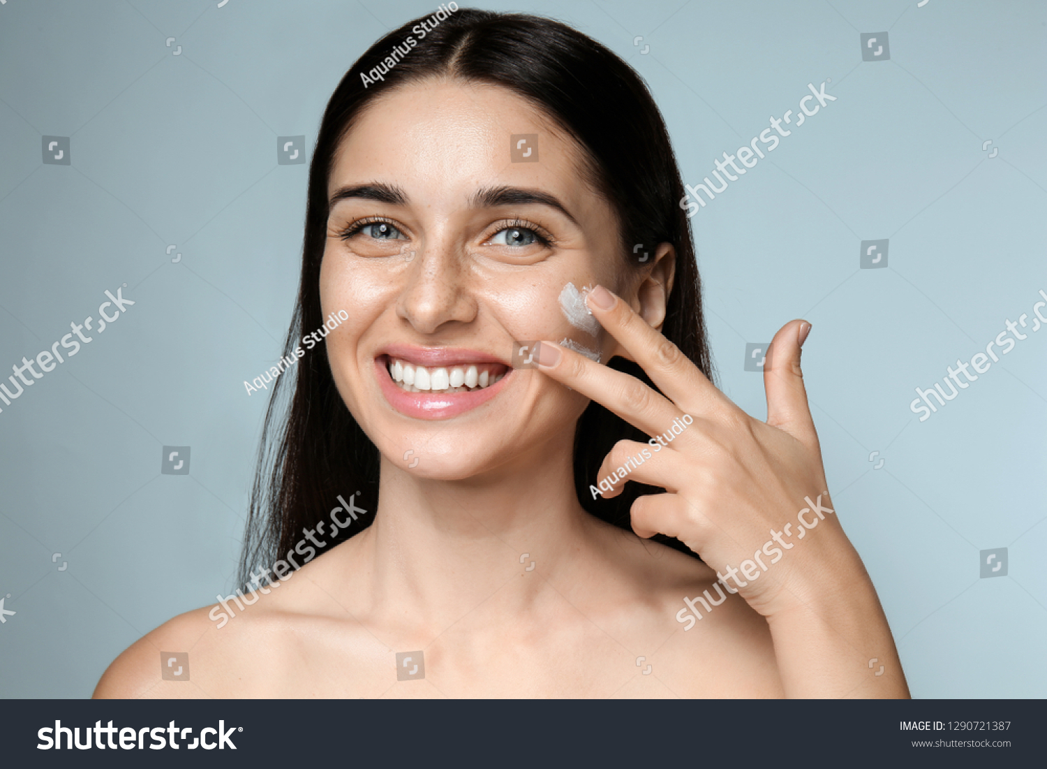 Portrait of beautiful young woman applying facial cream on color background #1290721387