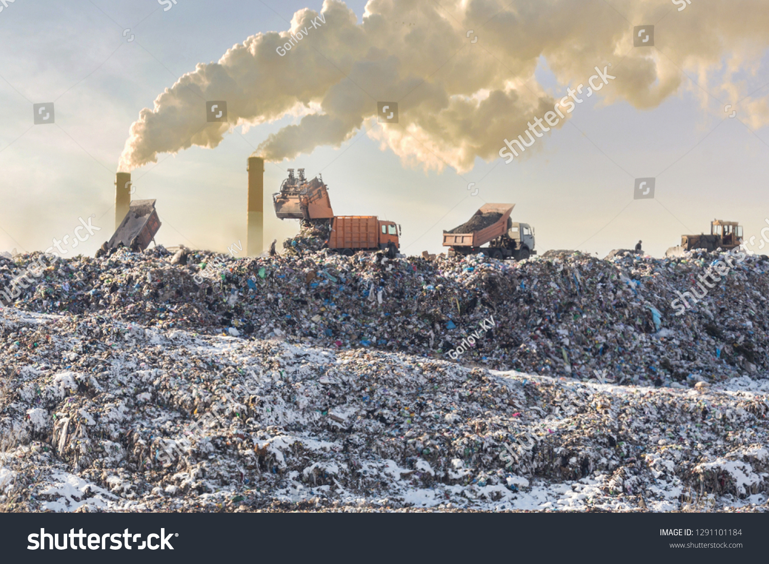 Dump trucks unloading garbage over vast landfill. Smoking industrial stacks on background. Environmental pollution. Outdated method of waste disposal. Survival of times past #1291101184