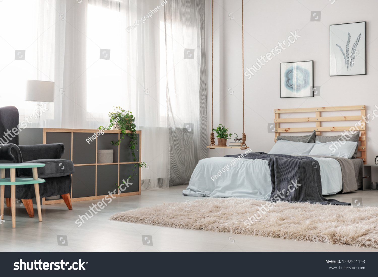 Fluffy white carpet on the floor of trendy bedroom with king size bed with wooden headboard, posters on the wall and fashionable swing as nightstand #1292541193