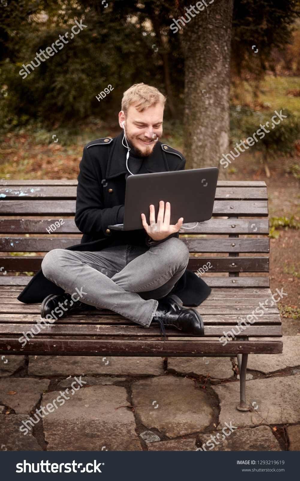 one young relaxed and smiling man, sitting casually on bench with crossed legs in public park, using laptop holding it with one hand. Formal wear or smart casual. Full length shot. #1293219619