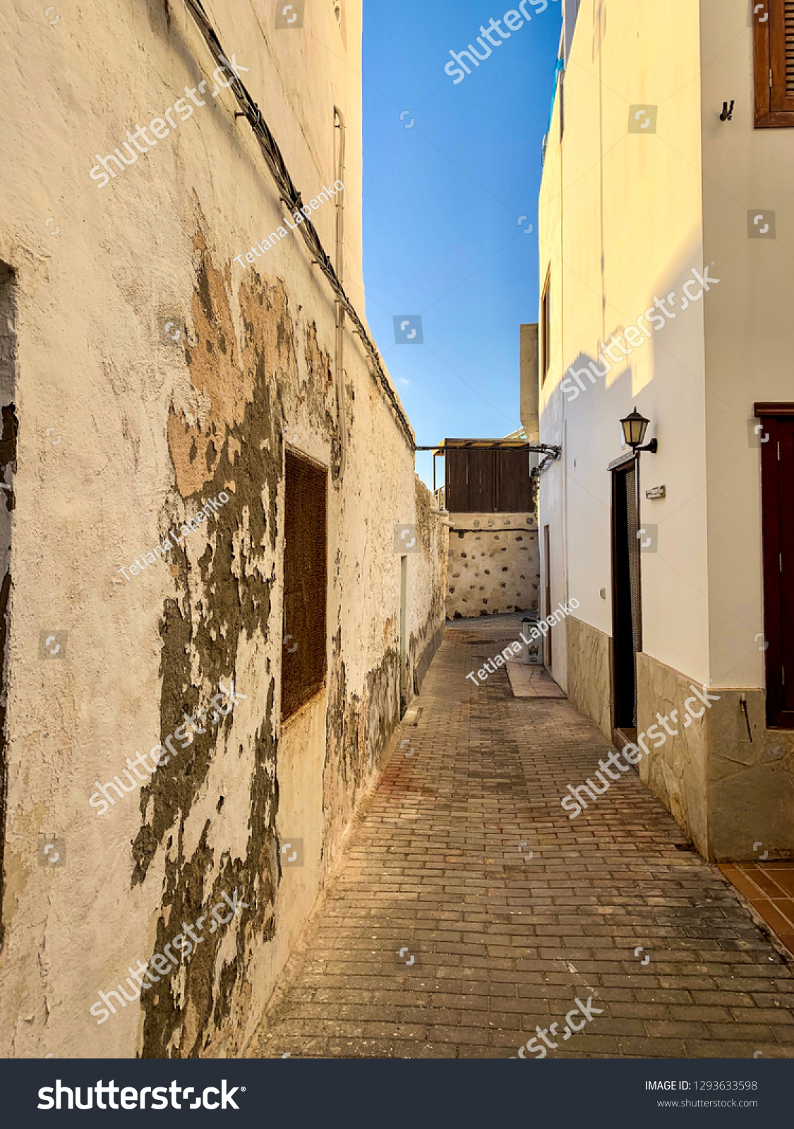 View of the street with white shabby walls of small buildings, architecture buildings with peeling paint, cosy street in fishing town El Cotillo on Atlantic coast  #1293633598