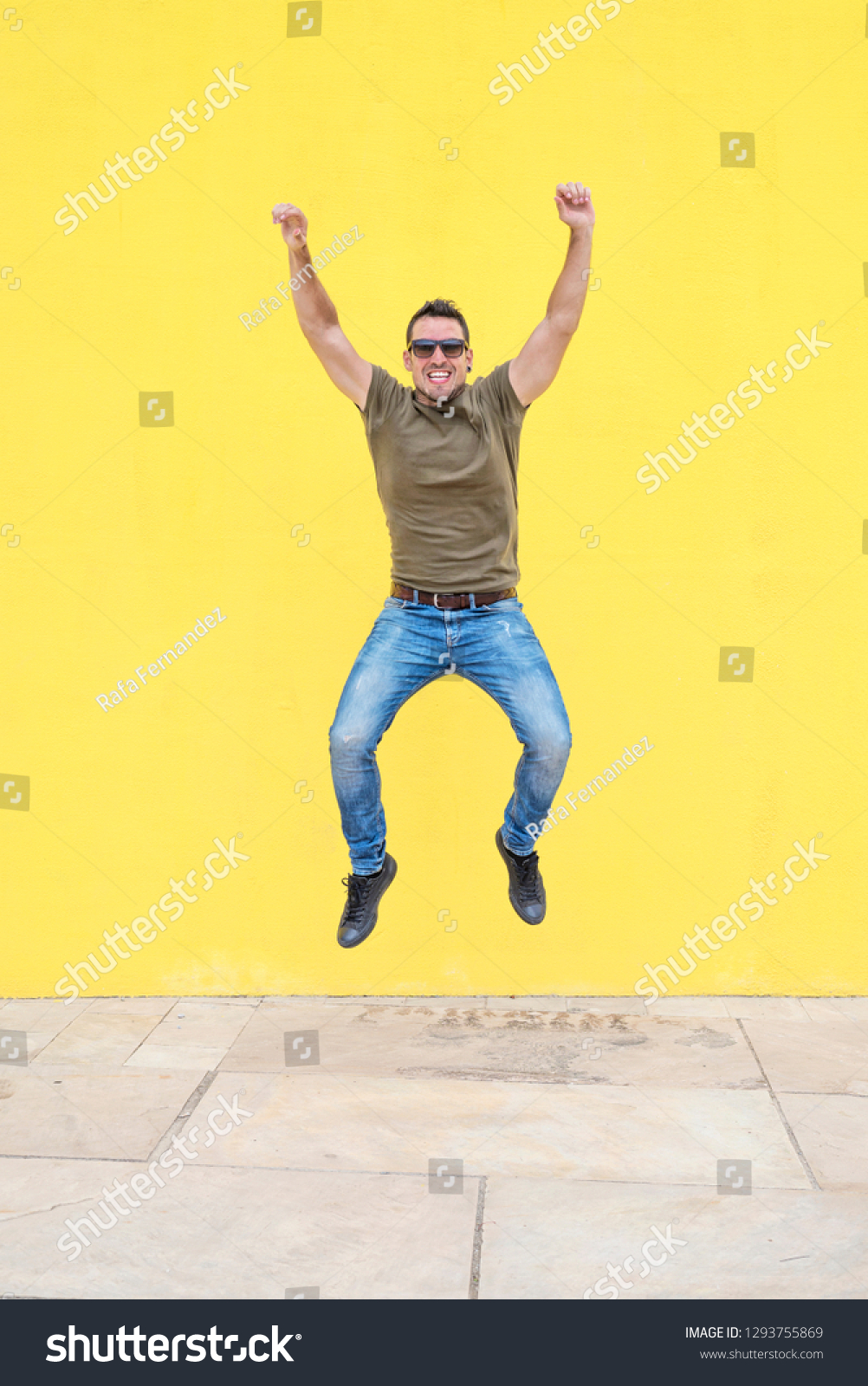 Front view of a young happy man wearing sunglasses jumping against a yellow bright wall in a sunny day #1293755869
