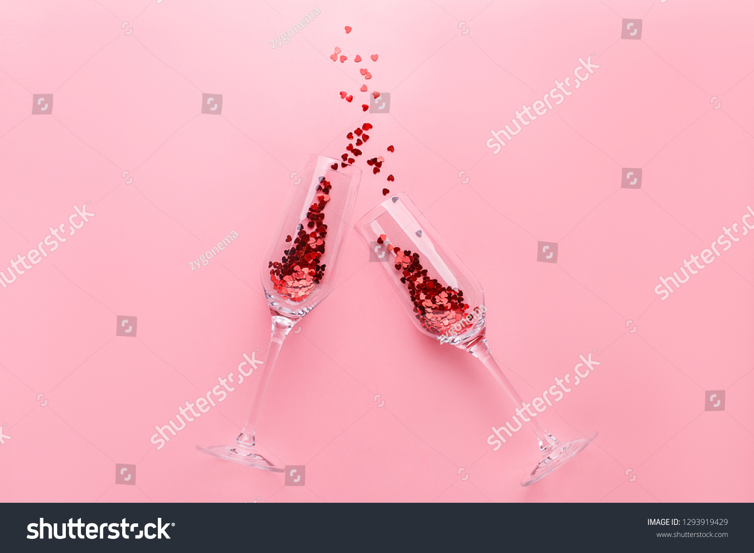 Two clinking champagne glasses with splash of red heart shaped confetti over pink background. Overhead view, copy space. Valentine's Day concept #1293919429