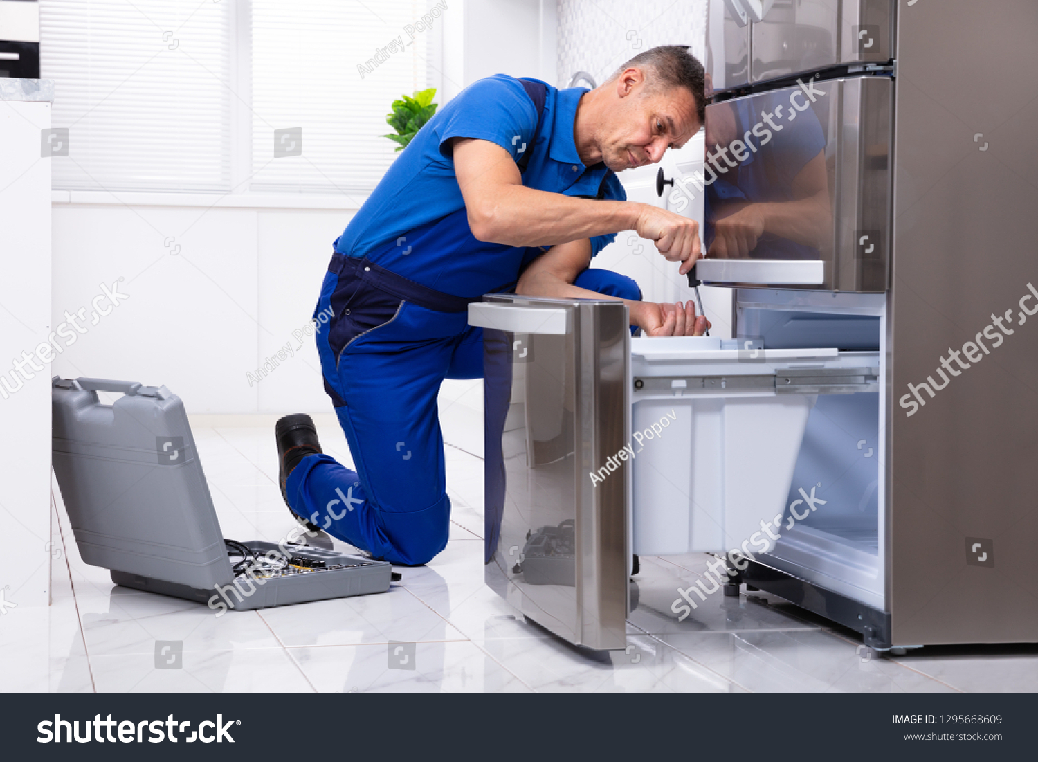 Mature Male Serviceman Repairing Refrigerator With Toolbox In  Kitchen #1295668609