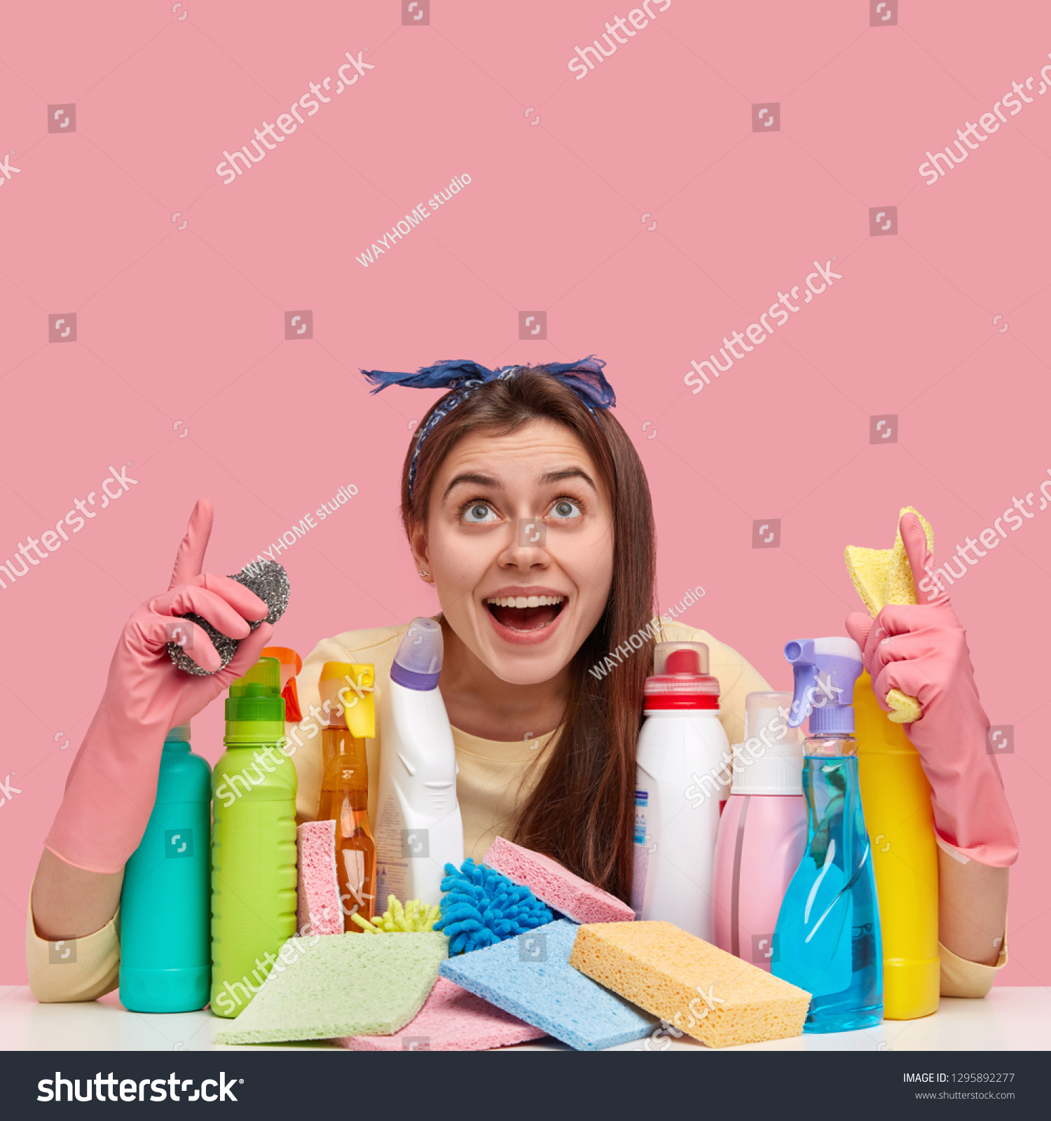 Vertical shot of good looking dark haired young woman with pleased expression, points at ceiling, wears headband, wears rubber protective gloves, ready for washing in house, uses cleaning agents #1295892277