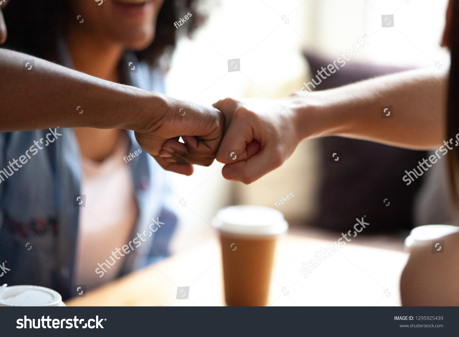 Close up multiracial best friends fist bumping, greeting each other, students celebrating successful exam results together, sitting in cafe, drinking coffee, symbol giving respect #1295925439
