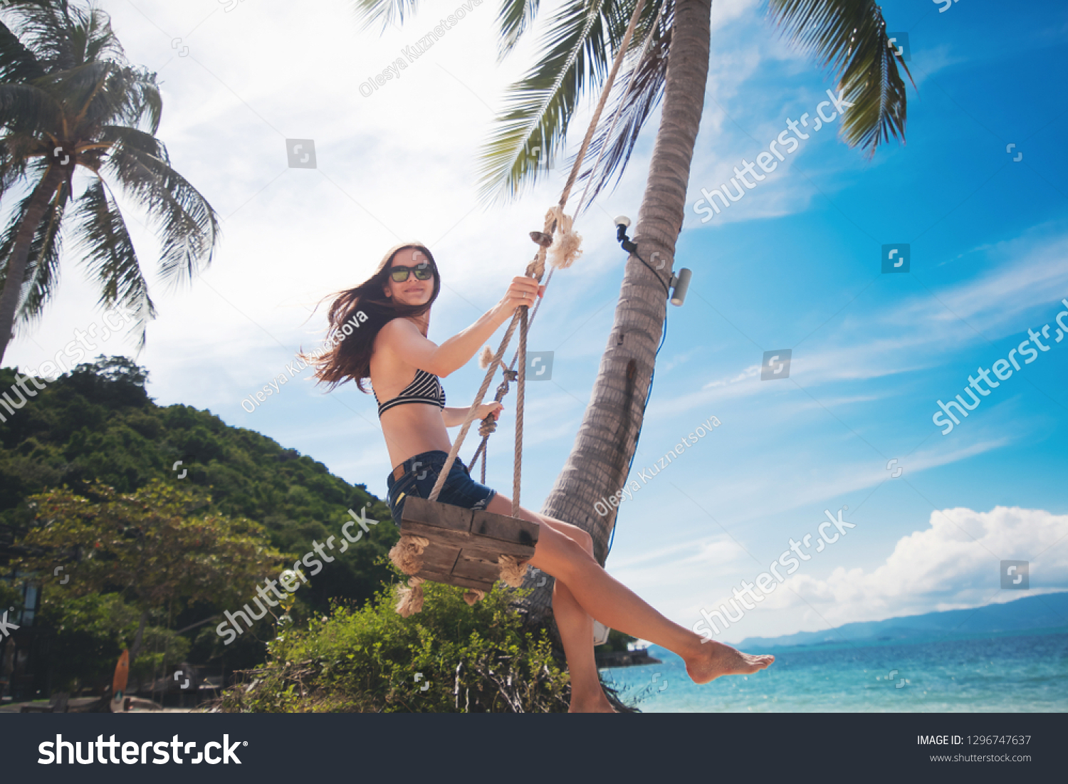 Young beautiful happy woman in top and shorts swinging on a swing on the shore of a tropical sea during vacation. Travel and vacation concept #1296747637