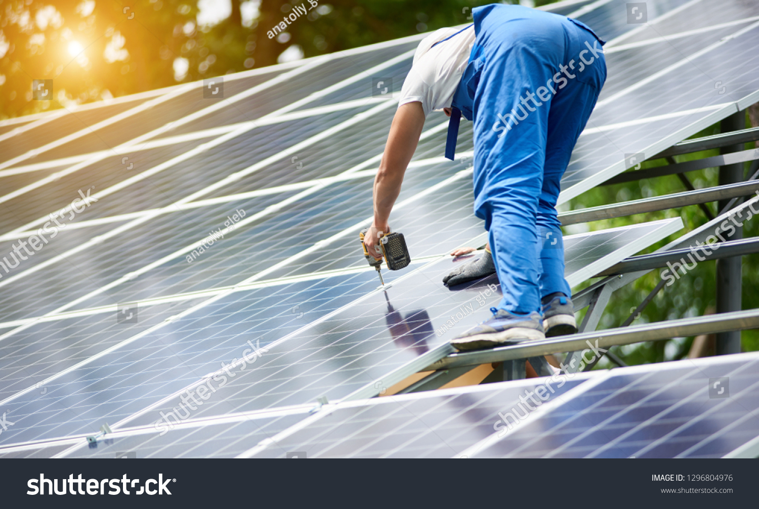 Construction worker connecting photo voltaic panel to solar system using screwdriver on shiny surface and lit by sun green tree background. Alternative energy and financial investment concept. #1296804976