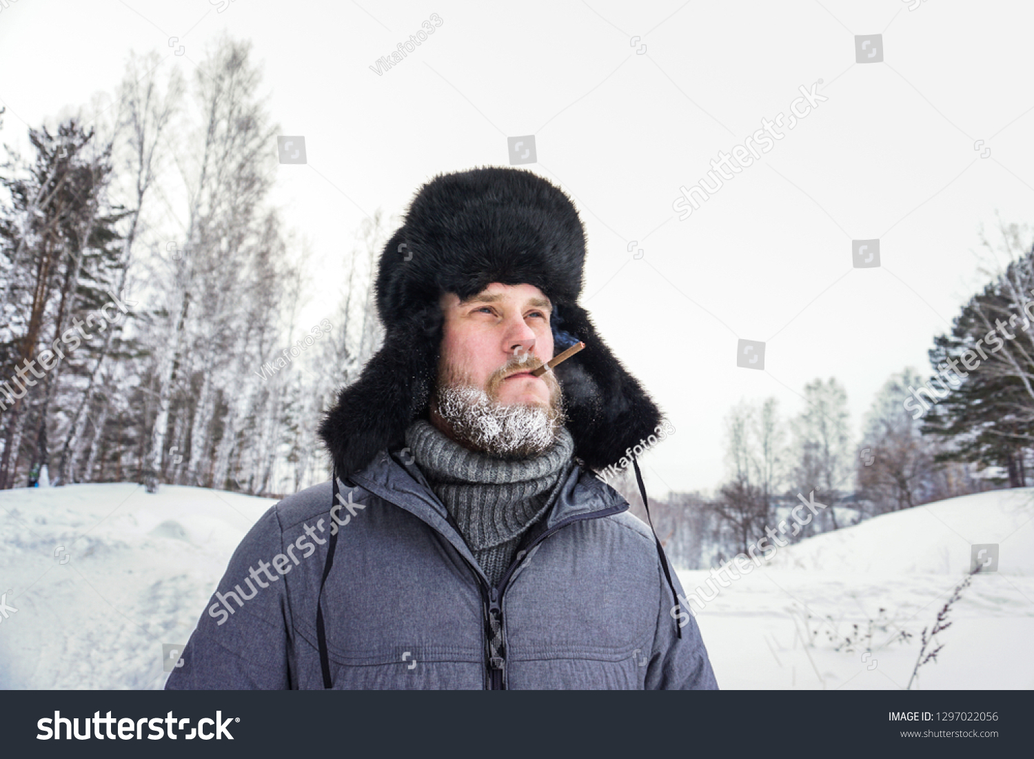 Siberian Russian man with a beard in hoarfrost in freezing cold in the winter freezes in a village in a snowdrift and wears a hat with a earflap smoking a cigarette #1297022056