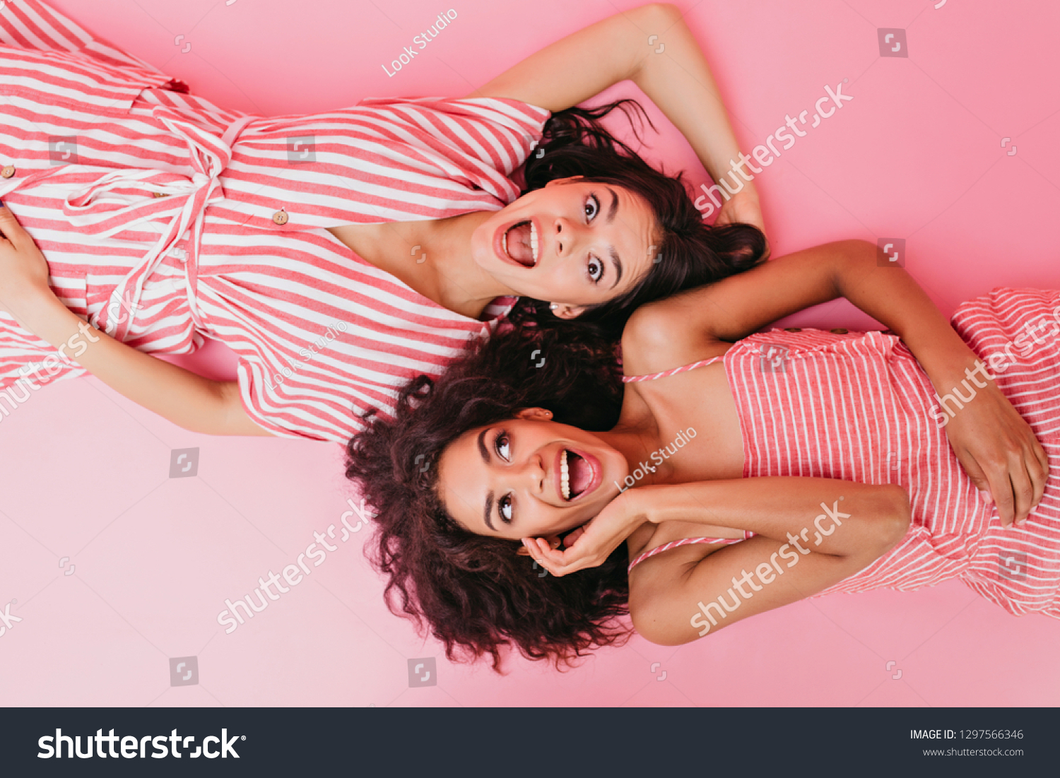 On isolated background two dark-skinned girls with beautiful hair and surprised faces pose for camera while lying on their backs #1297566346