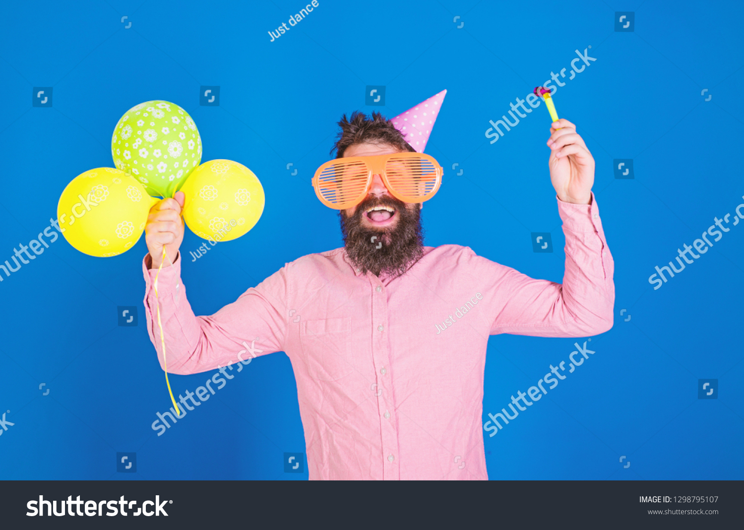 Hipster in giant sunglasses celebrating birthday. Man with beard and mustache on happy face holds party horn, blue background. Celebration concept. Guy in party hat with air balloons celebrates. #1298795107