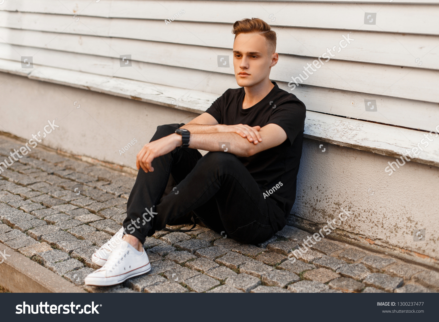 Handsome young man with a stylish hairstyle in stylish black summer clothes in fashionable white sneakers sits near a white historic building. Cool modern guy is relaxing. #1300237477