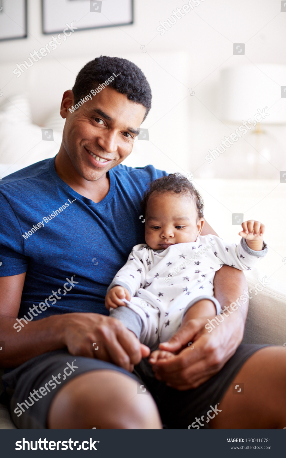 Close up of young adult African American  father sitting in an armchair holding his three month old baby son, smiling to camera, vertical #1300416781