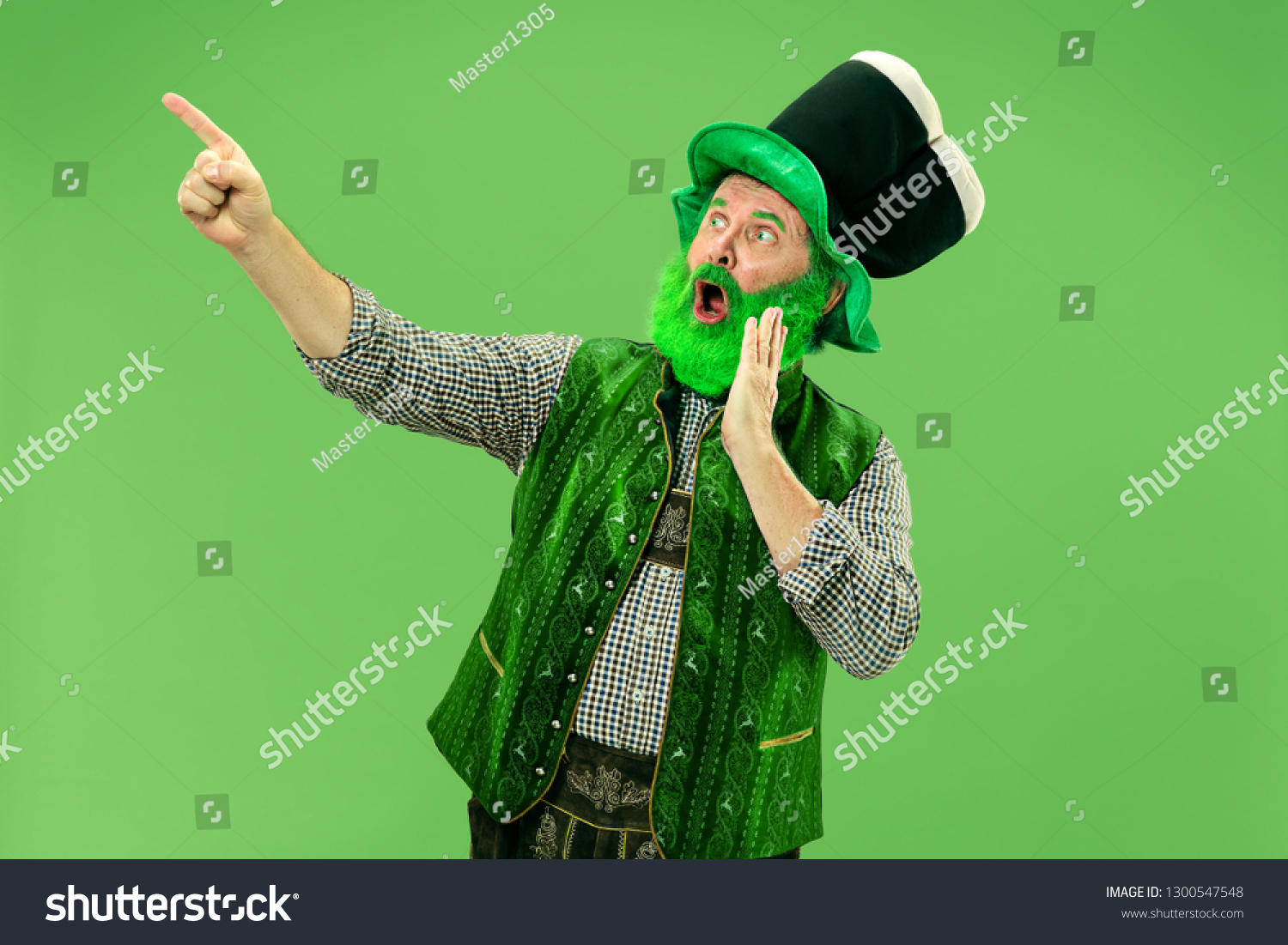 A smiling surprised happy senior man in a leprechaun hat with beard at green studio. He celebrates St. Patrick's Day. The celebration, festive, beer, holiday, alcohol, party, human emotions concept #1300547548